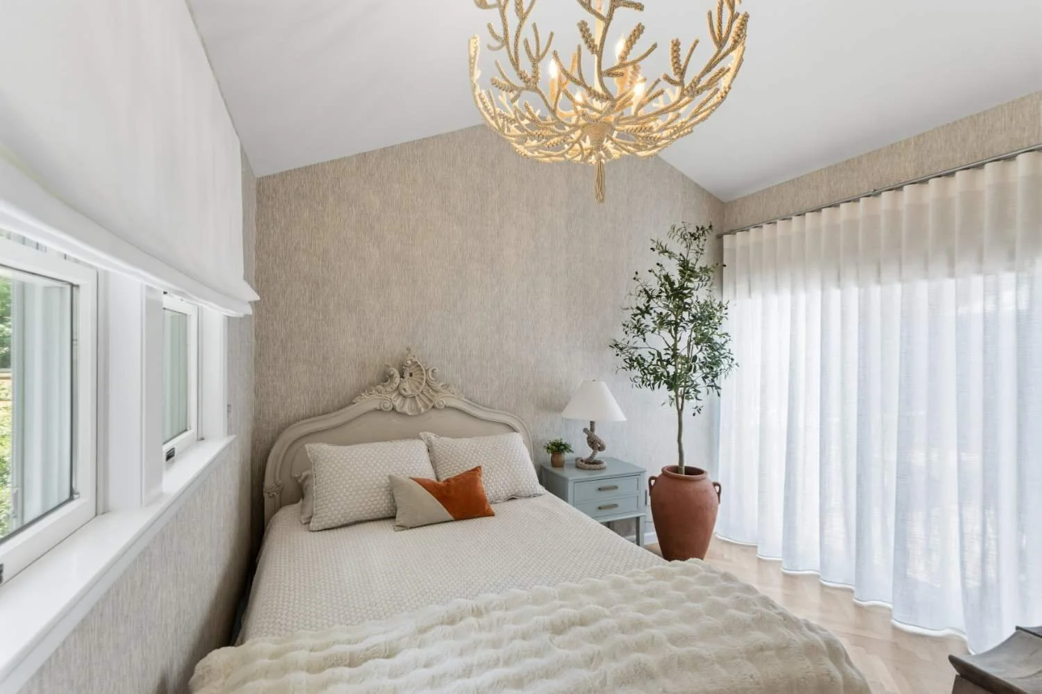 A bedroom with a large bed, decorative headboard, white bedding, throw pillows, a small bedside table with a lamp, a potted plant, and large windows with sheer curtains. There is a ceiling light fixture resembling a coral or tree branch.