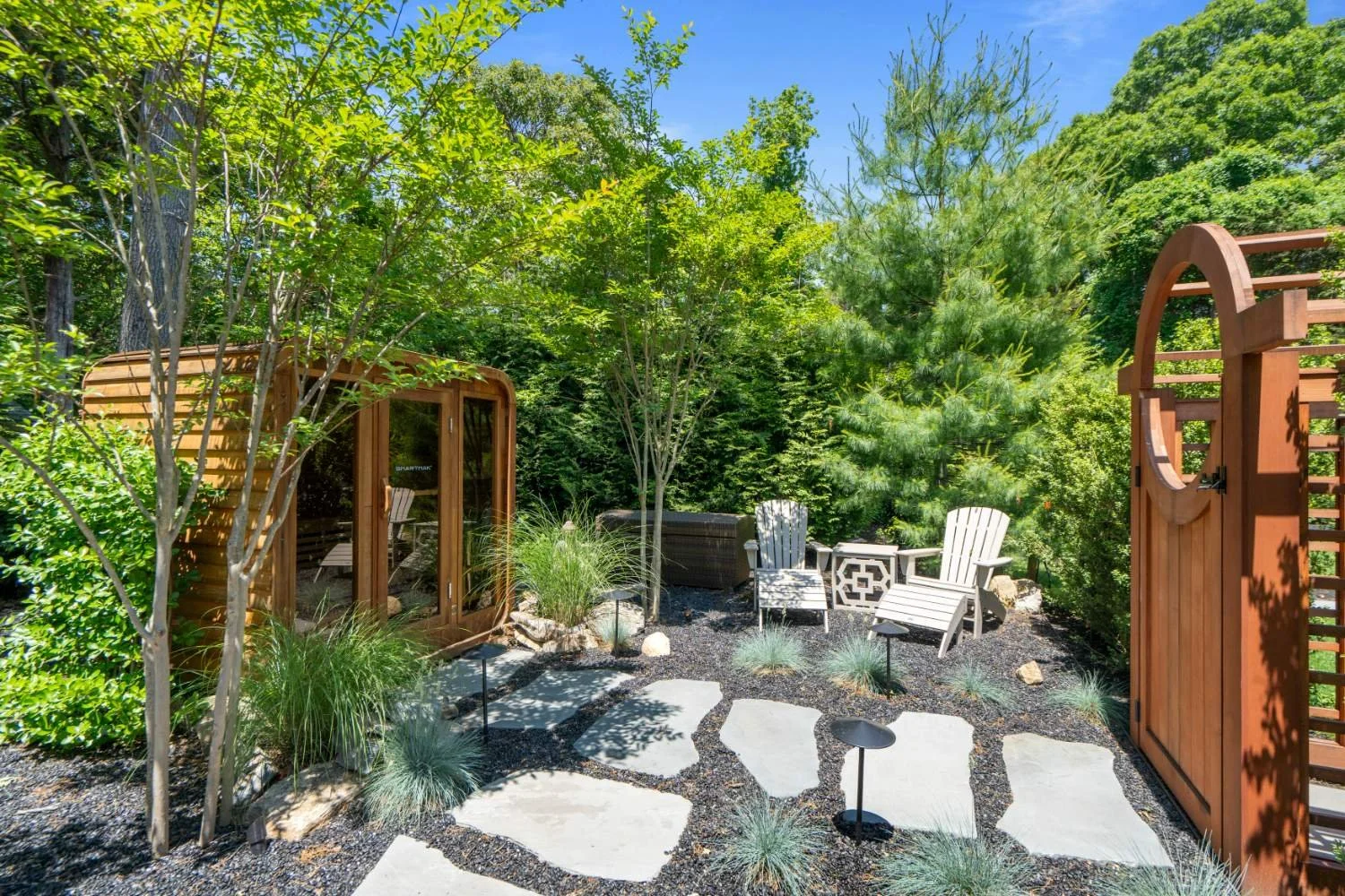 A backyard patio area with two white Adirondack chairs, a black wicker lounge chair, and decorative grasses, all surrounded by lush green trees and bushes under a clear blue sky.