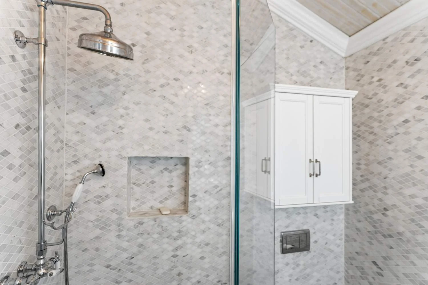 A bathroom with a shower area that has a large rain showerhead, a handheld shower, a wall niche with soap, and a glass partition. There is a white bathroom cabinet with silver handles on the wall outside the shower, and a ceiling with wooden panels.
