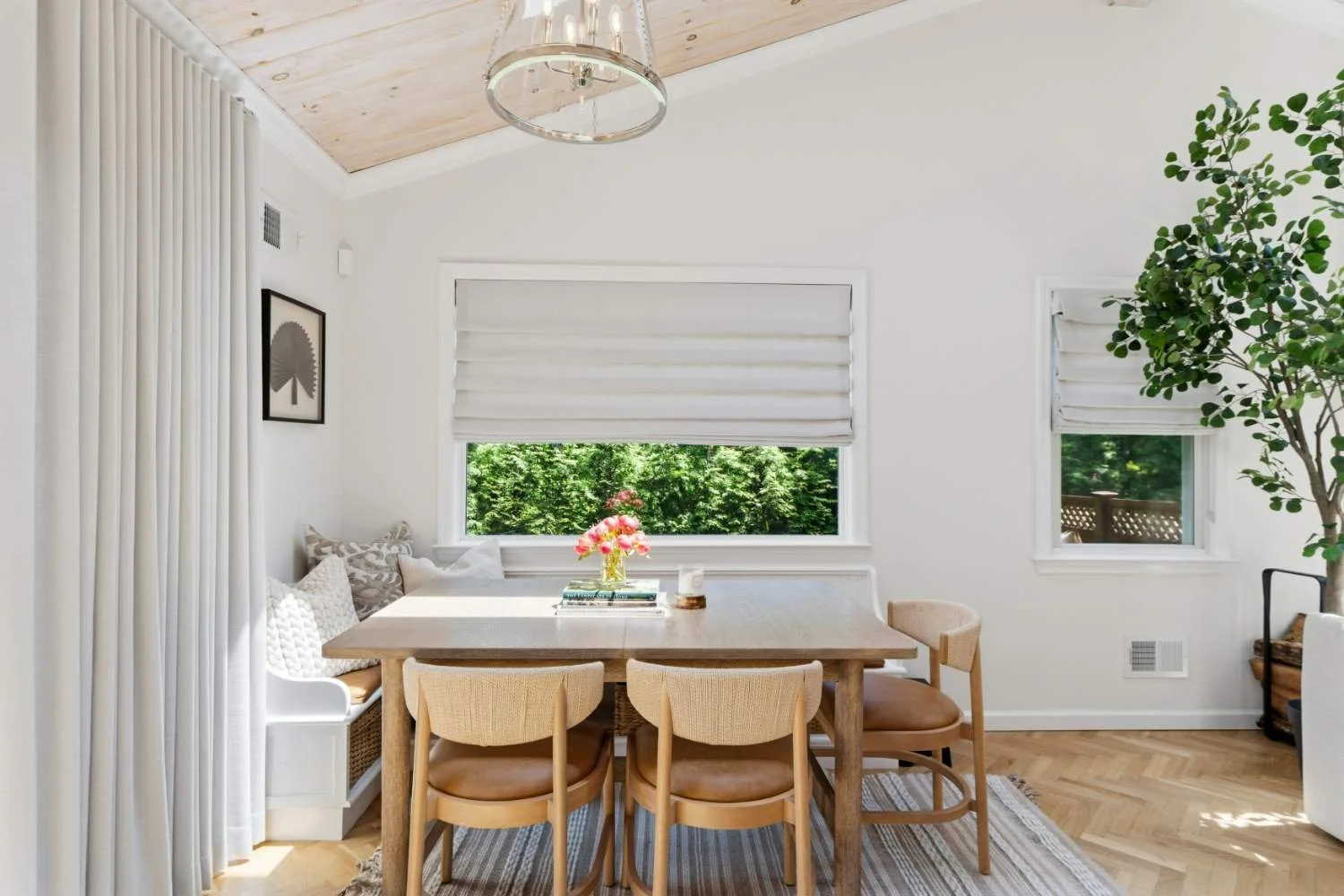 Bright dining room with a wooden table, beige chairs, and a window with white shades showing greenery outside. Decorative pillows on a built-in corner bench, a small vase with pink flowers, and a large potted plant in the corner.