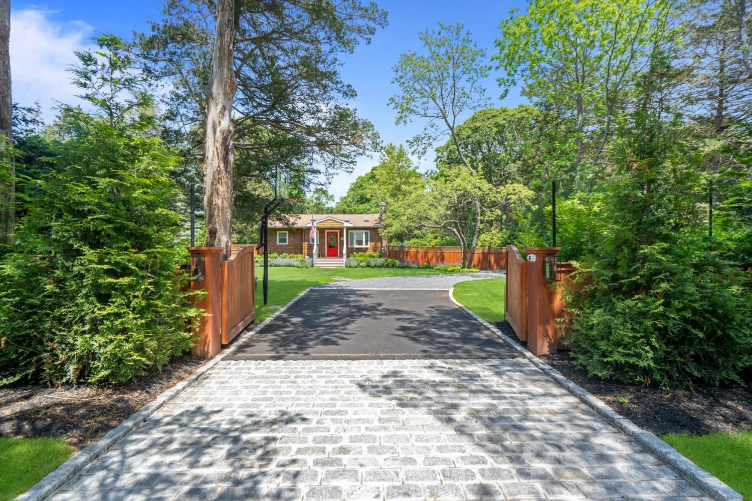 View of a house with a red door, seen through an open wooden gate with black accents, surrounded by lush green trees and bushes on a sunny day.