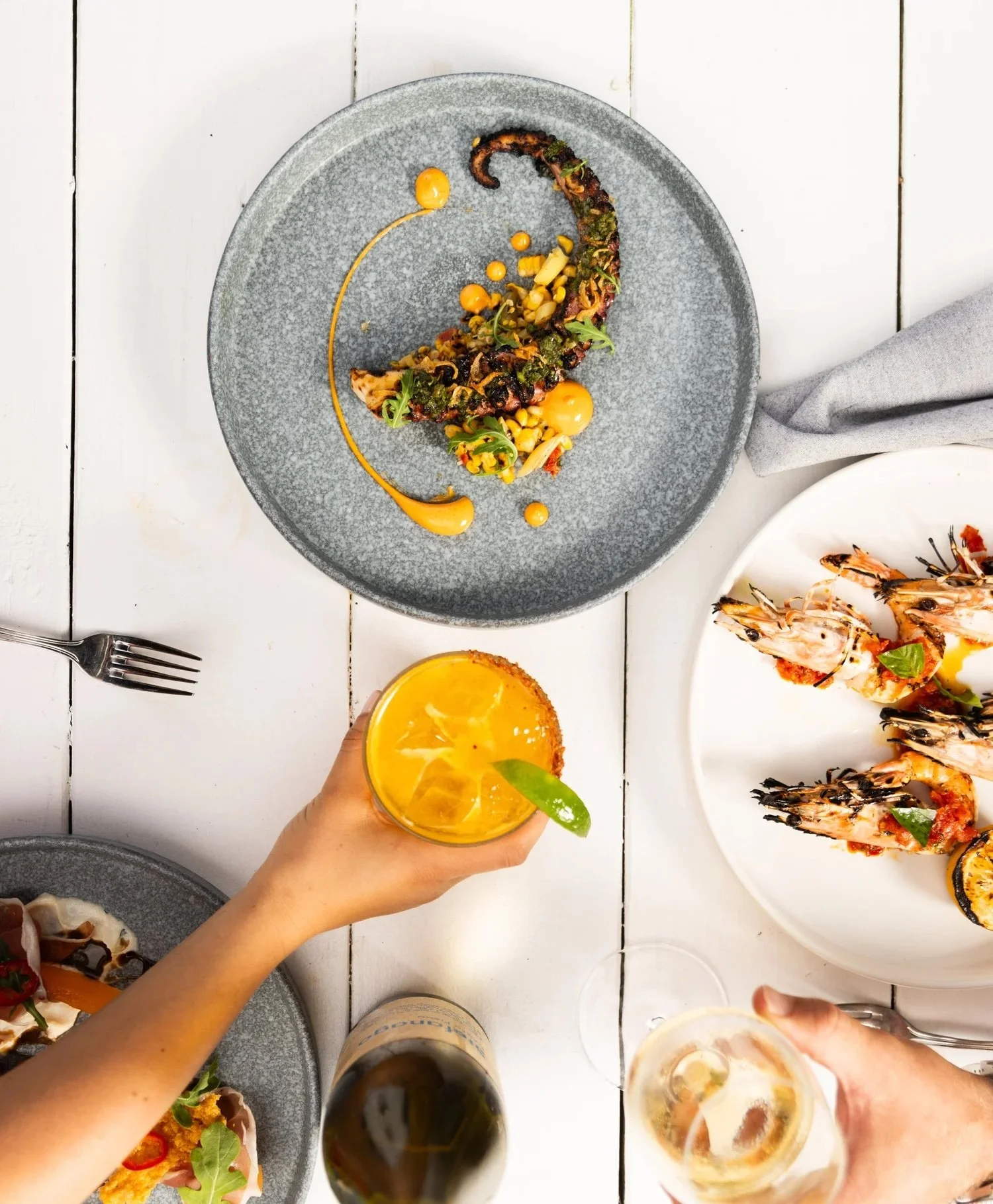 People holding glasses of orange-colored drink at dining table with various plates of seafood and food, including grilled octopus with vegetables, shrimp, and other dishes.