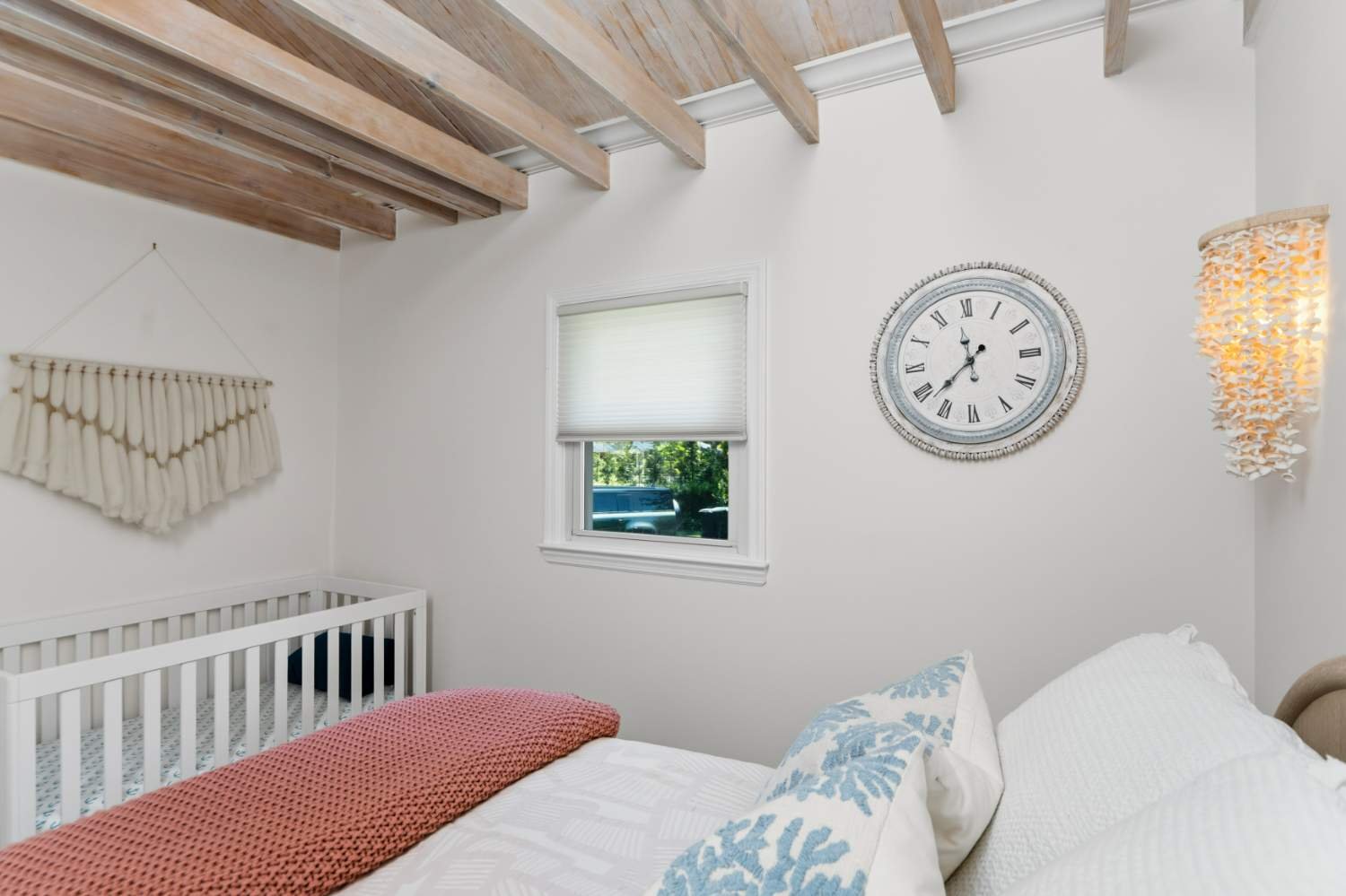 A bedroom with white walls and a wooden ceiling beam, a window with a white blind showing greenery outside, a wall clock with Roman numerals, a woven wall hanging, a bed with white linens, pillows with blue patterns, and a coral-colored blanket.