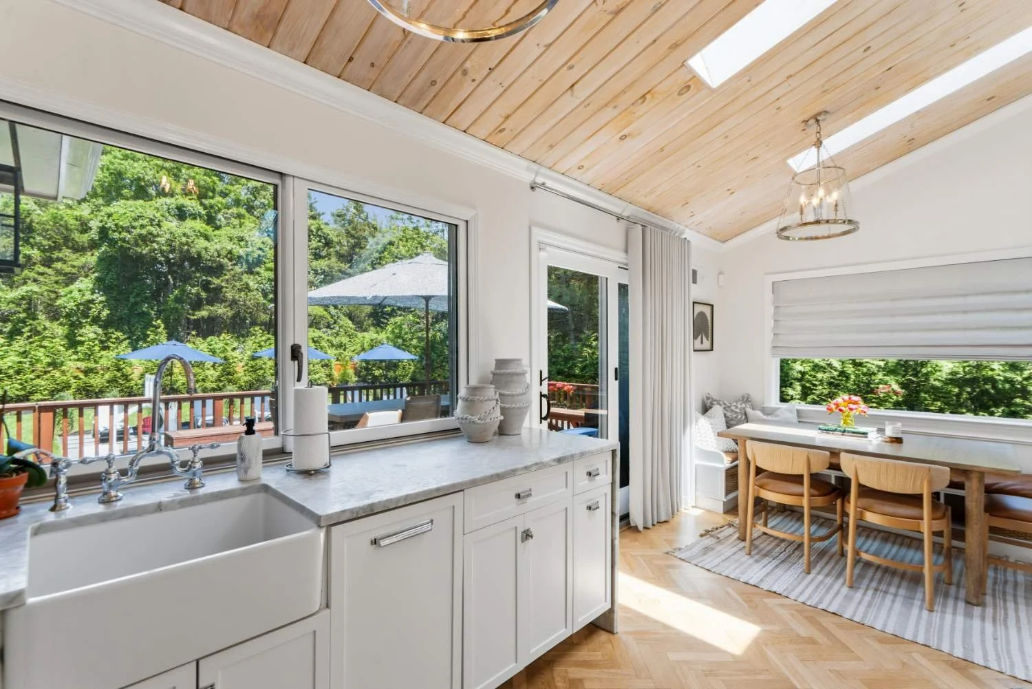 Bright kitchen with large windows showing outdoor patio with umbrellas, a wooden dining table with chairs, and a light wood-paneled ceiling.