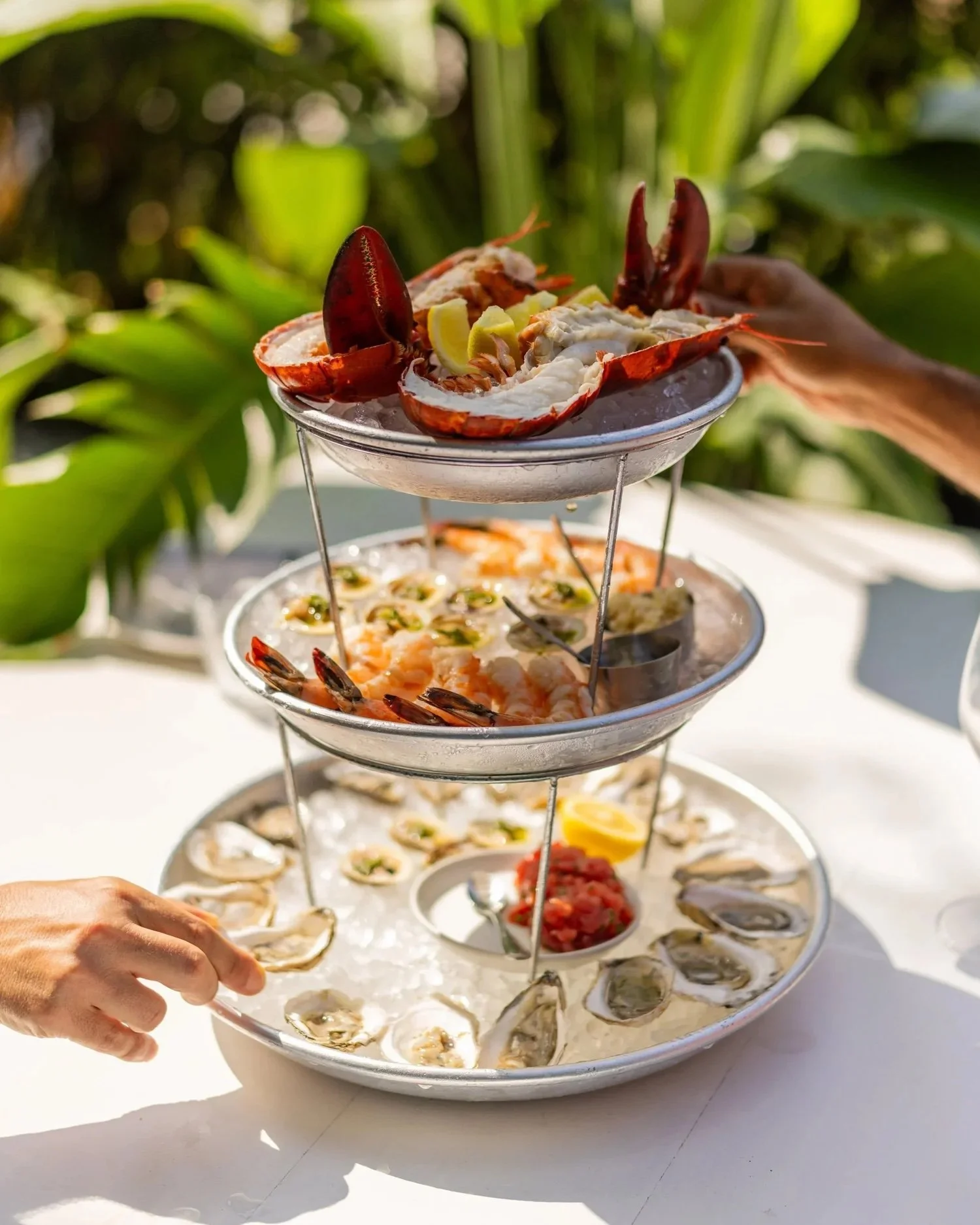 A multi-tiered tray of seafood, including oysters, shrimp, and lobster, garnished with lemon wedges on a white table outdoors.