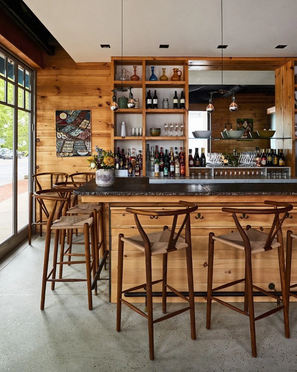 Interior of a modern bar or cafe with wooden decor, bar stools, bottles, glasses, and decorative vases on shelves.