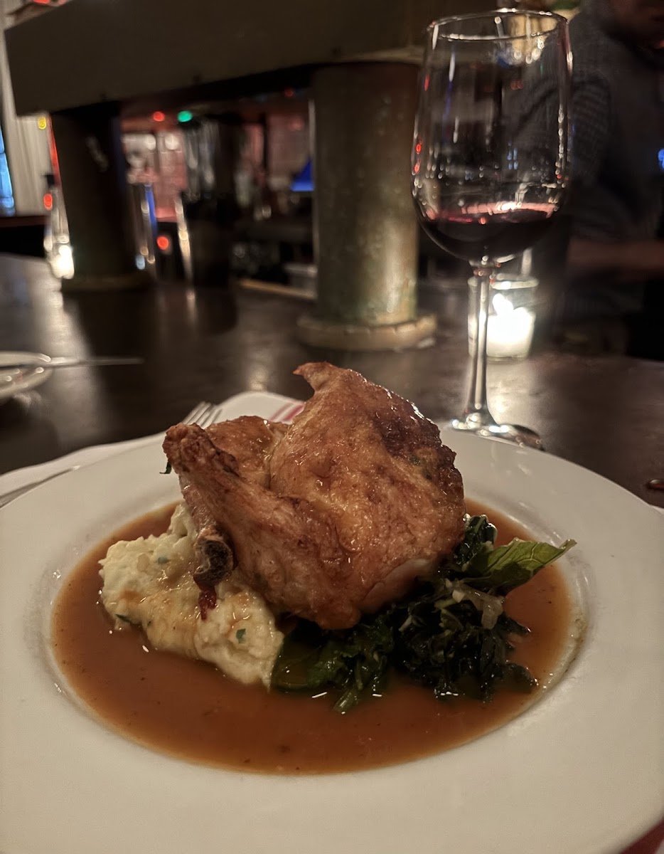 A plate with roasted chicken, mashed potatoes, and cooked greens in gravy, with a glass of red wine in the background at a restaurant.
