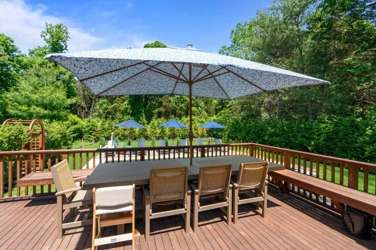 Outdoor wooden deck with large patio table, chairs, and umbrellas surrounded by green trees under a bright blue sky.