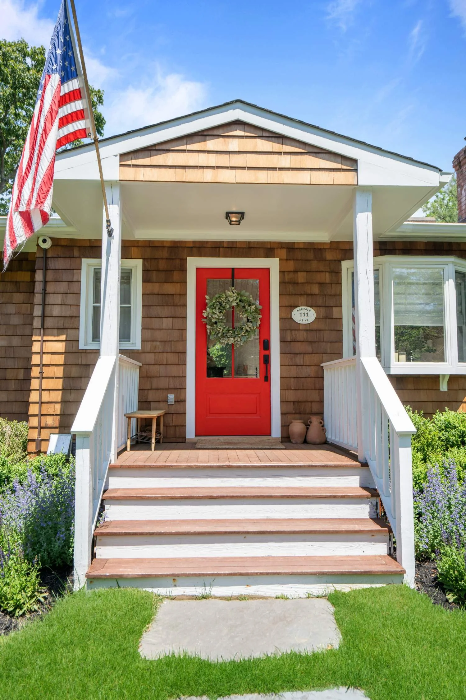 Front porch of a house with a red door decorated with a wreath, white porch railings, steps, a small bench, potted plants, and an American flag on a pole. The house has brown wooden siding, a window on each side, and a small oval sign with the addres
