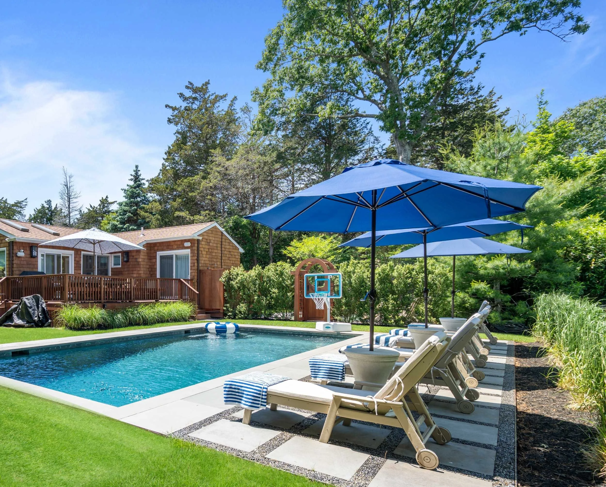 Backyard with swimming pool, lounge chairs with blue striped towels, blue patio umbrellas, green trees, and a small house with a wooden deck.