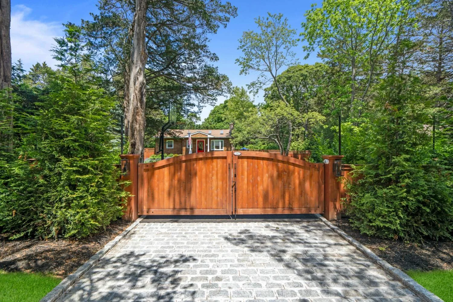 A wooden gate at the end of a cobblestone driveway, with tall greenery and trees on either side, and a house visible in the background.
