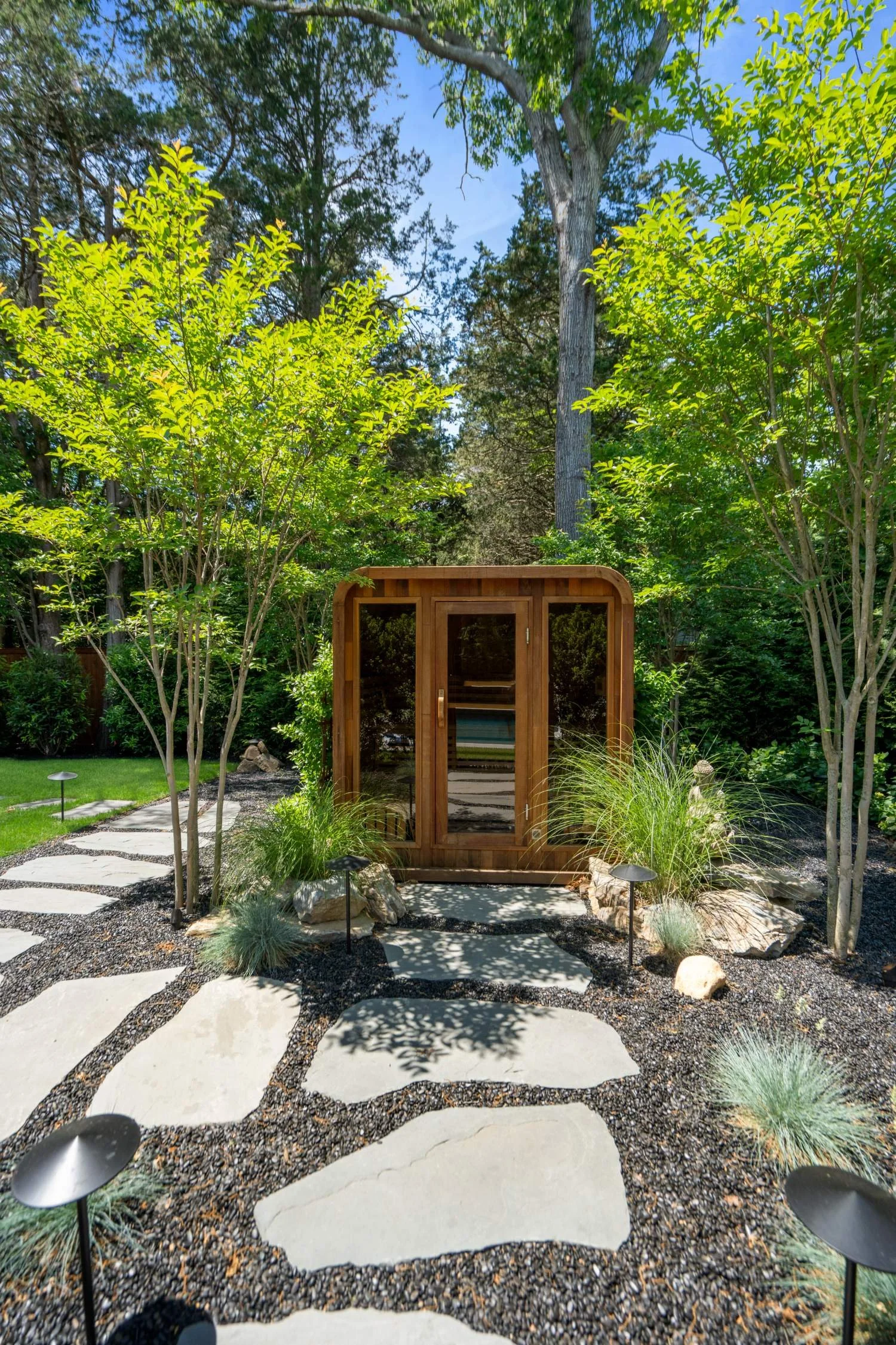 A landscaped backyard with a stone pathway, a small wooden shed, green trees and shrubs, and small outdoor lights.