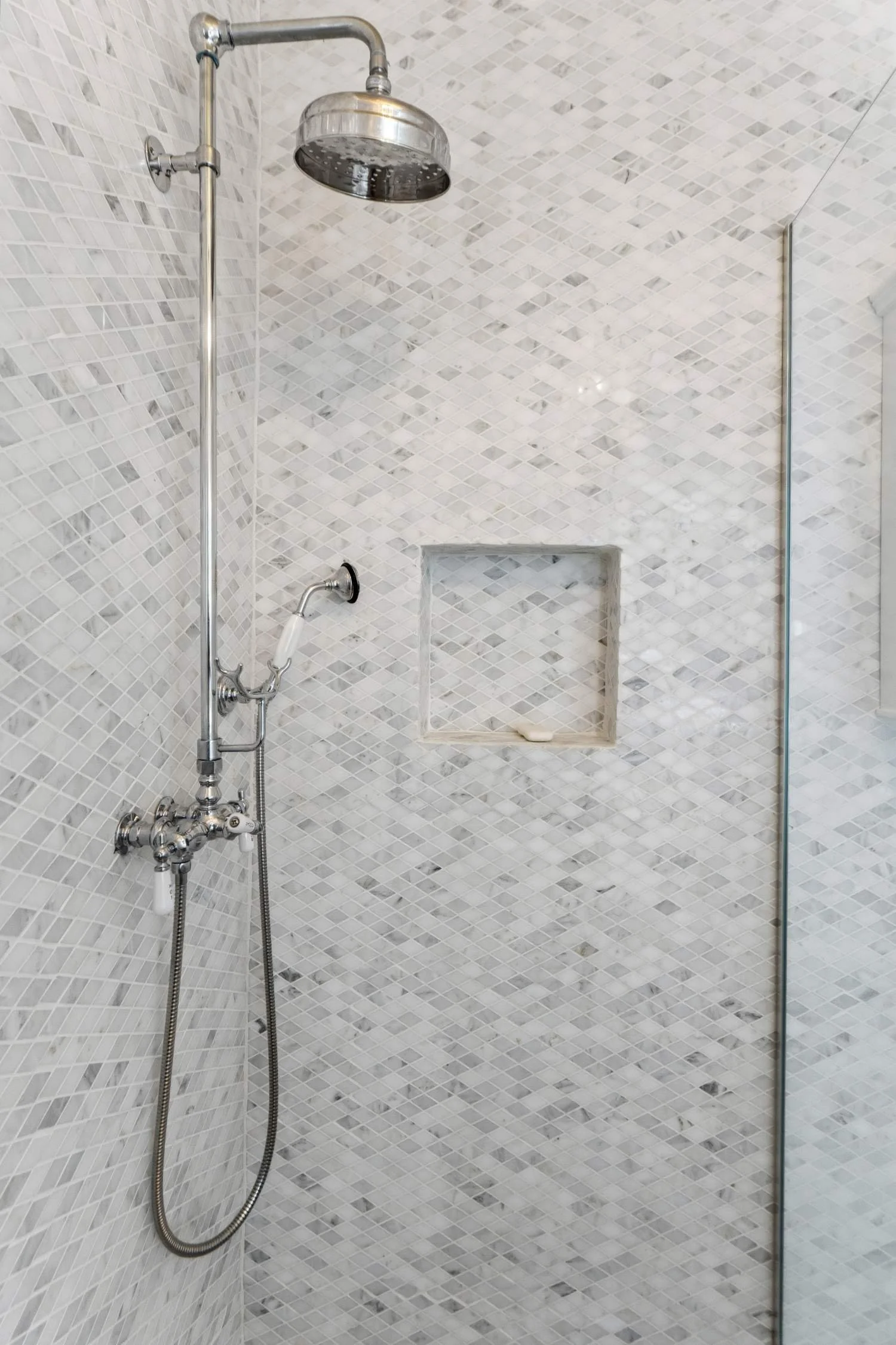 A modern shower with a rain shower head, a handheld shower, and a built-in tiled niche for toiletries. The walls are covered in small, white, square mosaic tiles.