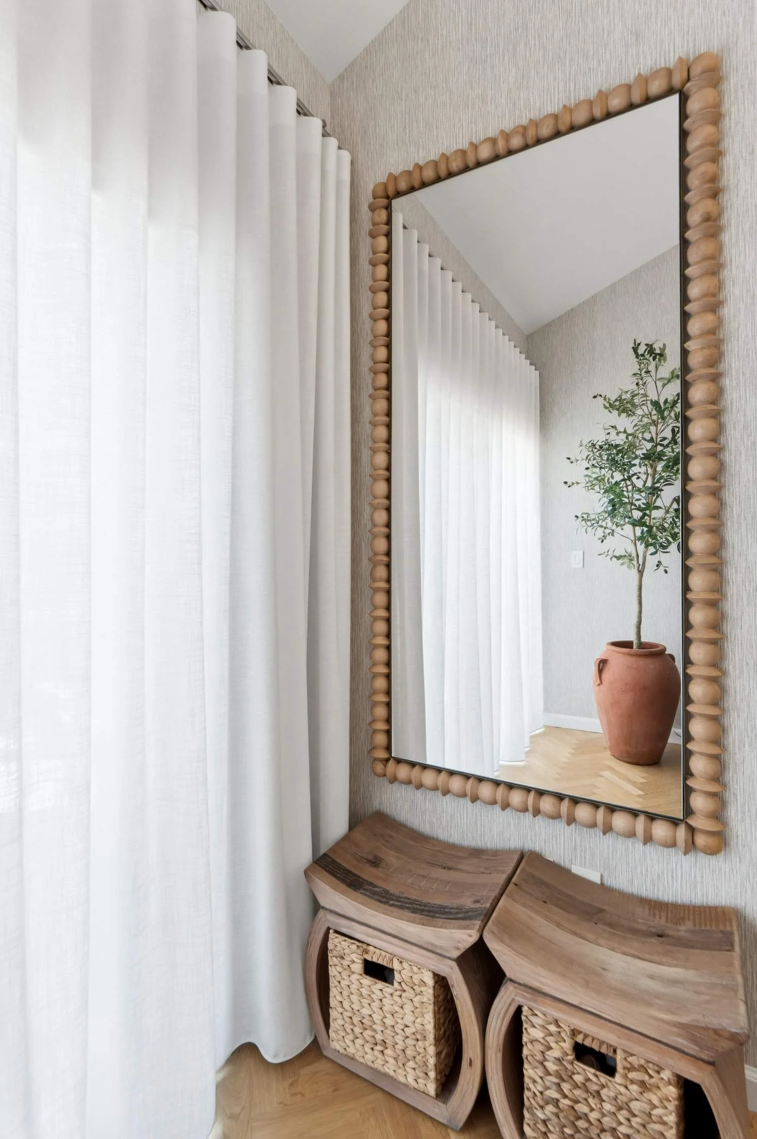 A mirror with a wooden bead frame hangs on a wall next to white curtains. The mirror reflects a potted plant and a corner of the room. There is a wooden bench with rounded edges and woven baskets underneath it.