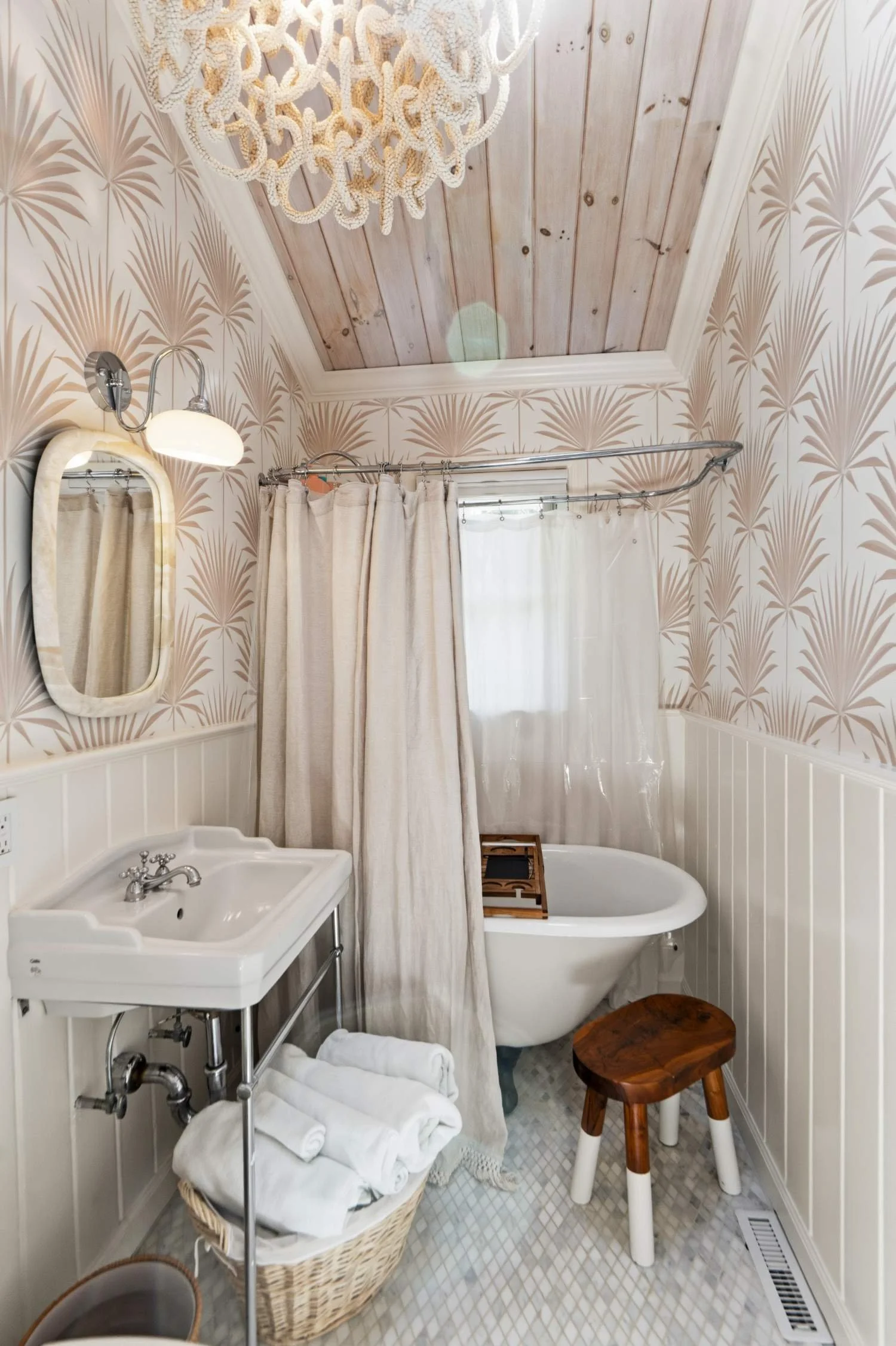 A cozy bathroom with patterned wallpaper, a wooden ceiling, a vintage-style bathtub, a white sink with a mirror, and a wicker basket with folded towels. There is a small wooden stool, a ladder for towels, and curtains covering the window and shower a