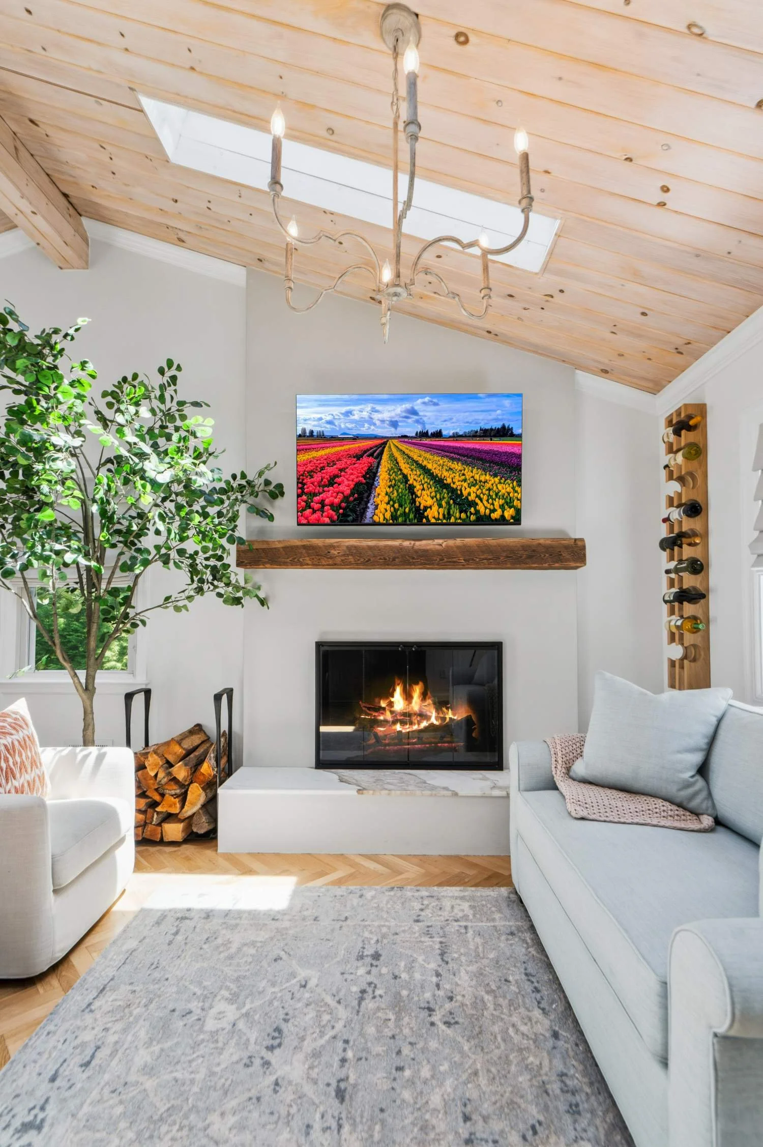 Living room with white sofa, fireplace, large window, potted plant, wooden ceiling with skylights, chandelier, wall-mounted TV displaying colorful tulip fields, wooden log holder, and a gray rug.