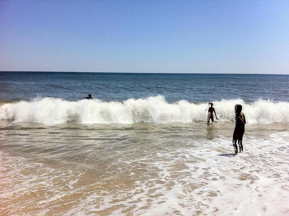 People enjoying the ocean waves at the beach, some standing in the shallow water and one swimming farther out, under a clear blue sky.