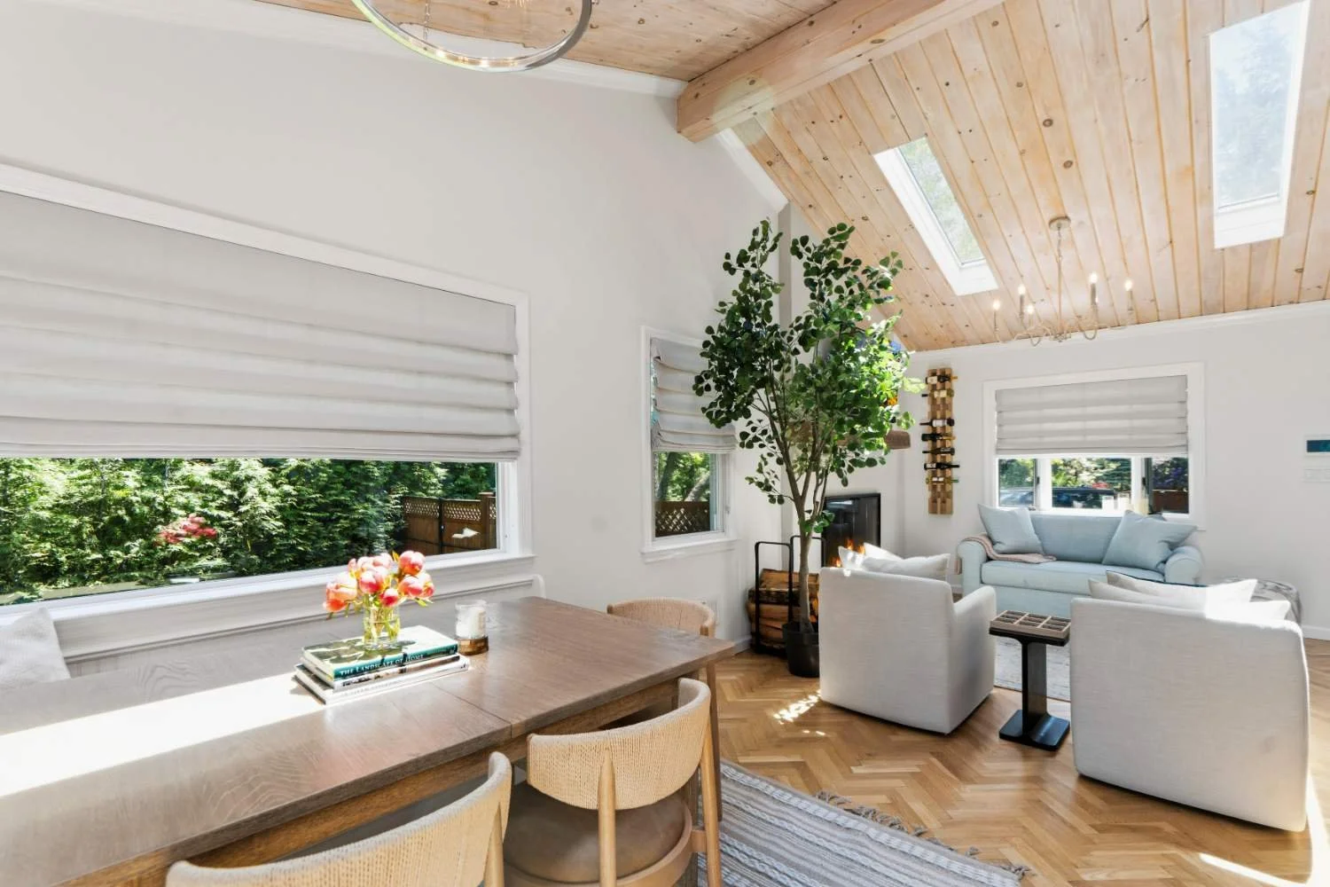 Bright living room with large windows, white walls, wooden ceiling, and hardwood floor. Features a dining table with chairs, a potted plant, and a cozy seating area with white and light blue sofas and armchairs.