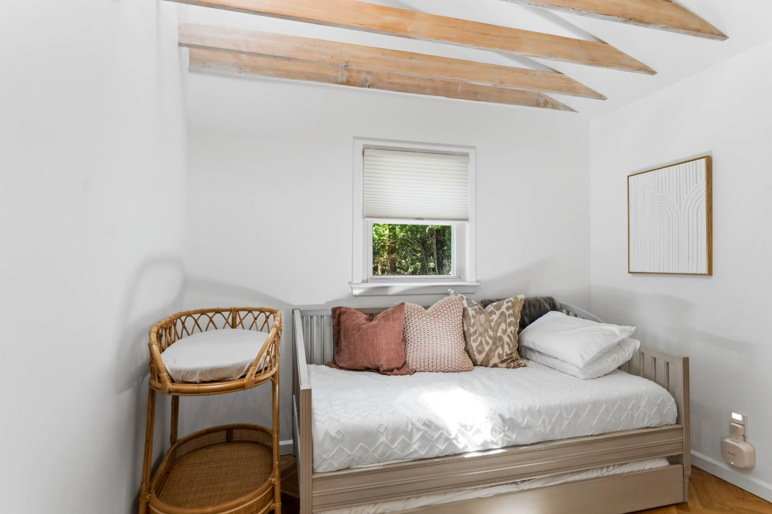 A bedroom with a white bed, multiple pillows, a window with a white blind, and wooden ceiling beams.