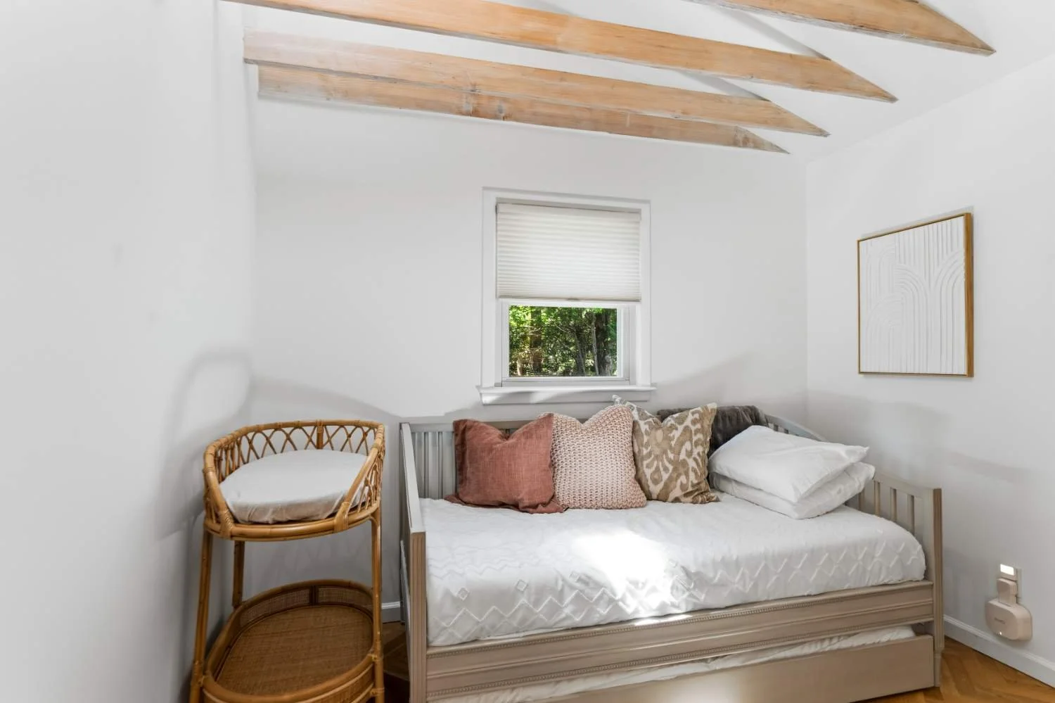 A bedroom with white walls, a window with a white blind, a bed with an assortment of pillows, and a wicker side table in the corner.