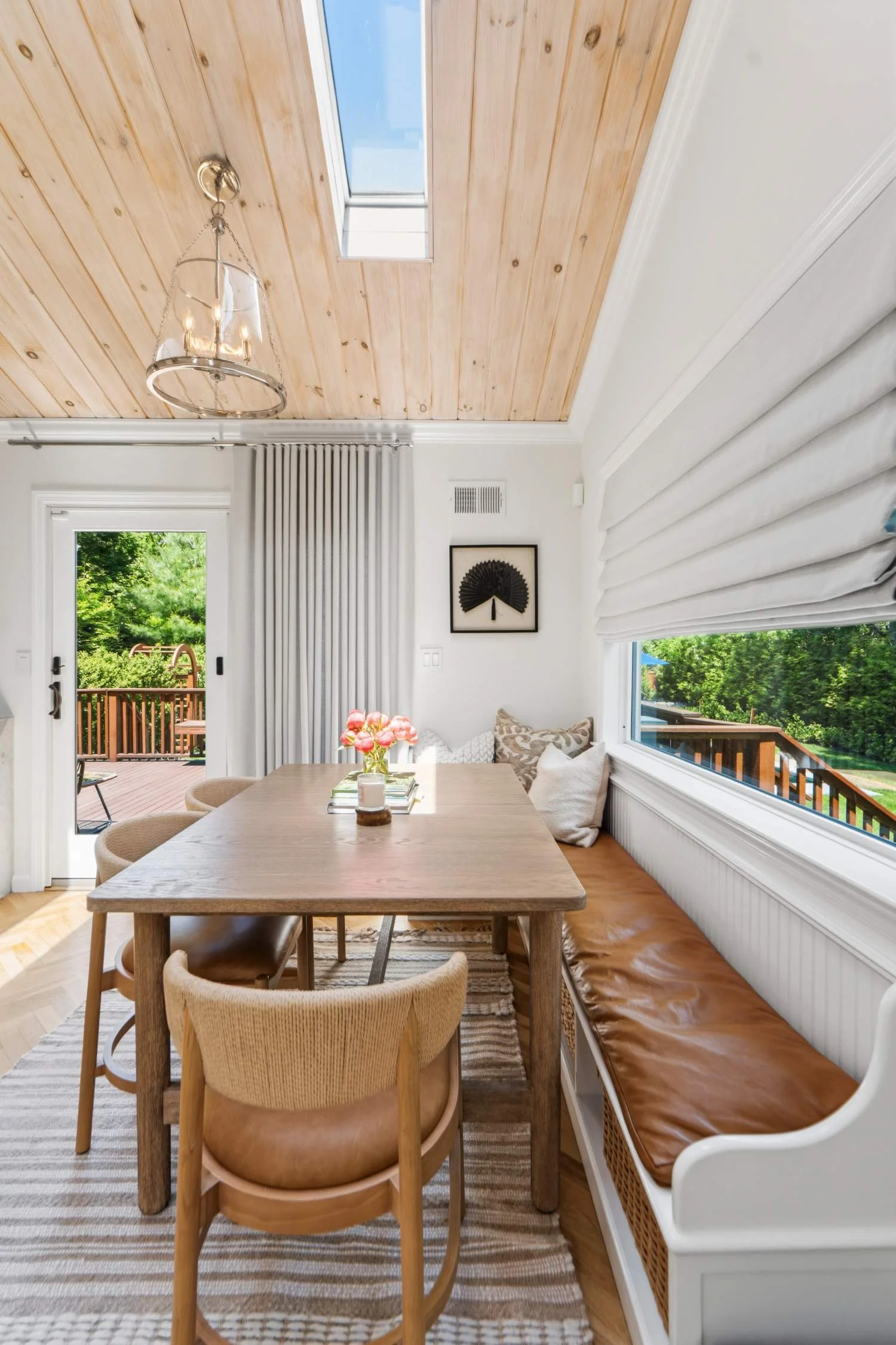 A dining room with a wooden table and chairs, a bench with cushions, large windows with white blinds, and a glass door leading to an outdoor deck with trees in the background.
