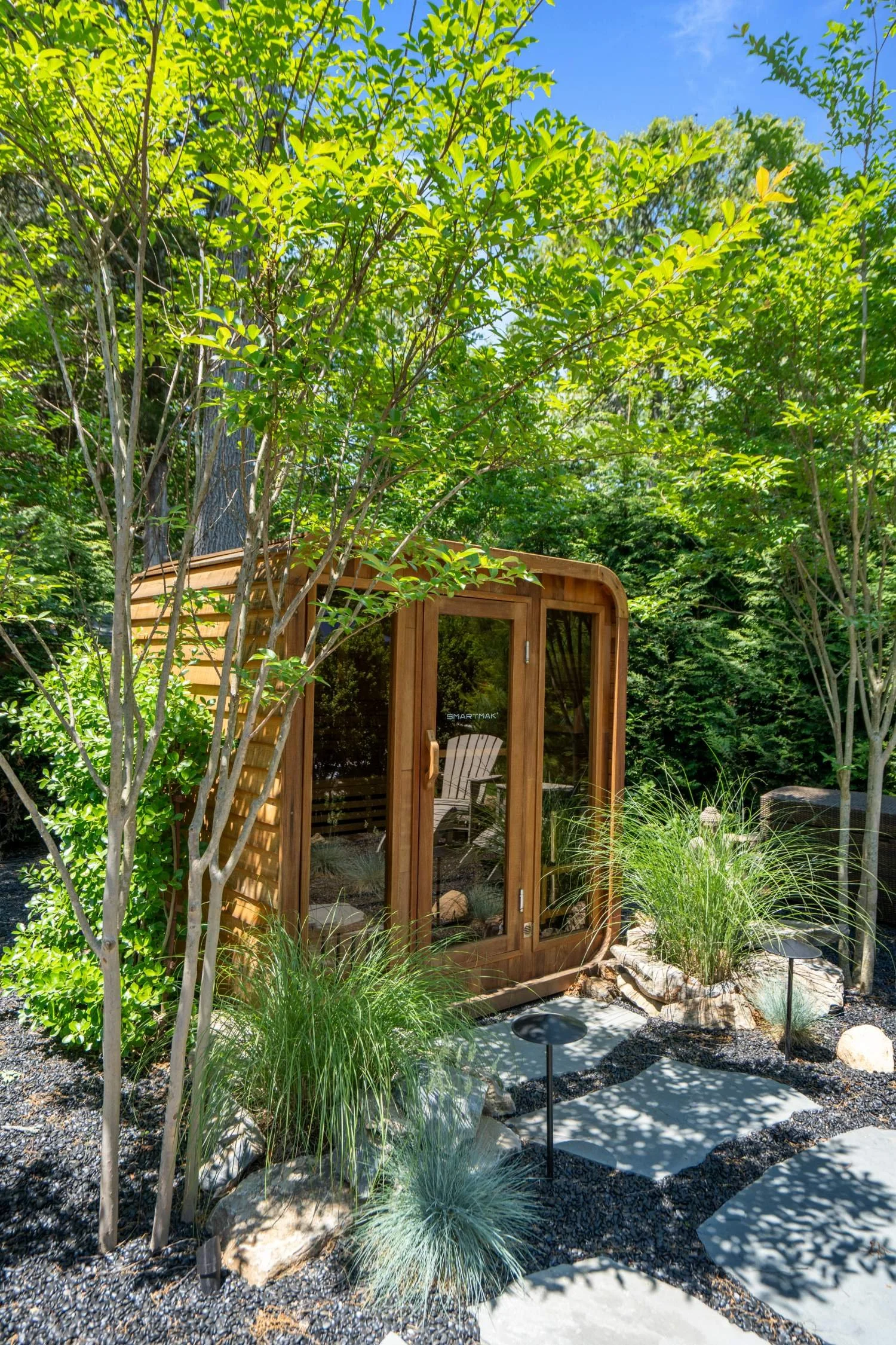 A small wooden garden shed surrounded by lush green trees and plants, with a curved window and door, and a landscaped yard with rocks, gravel, and pathway lighting.