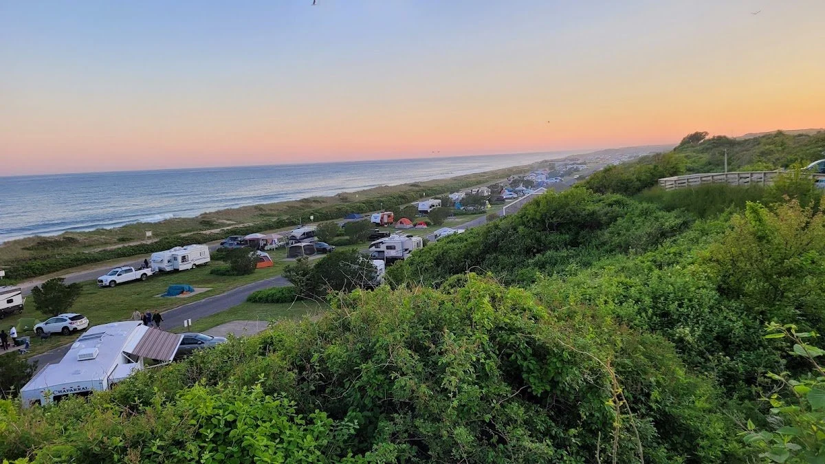 View of a beachside camping area at sunset, with tents, cars, and RVs lined up on grassy land near the ocean and green shrubbery in the foreground.