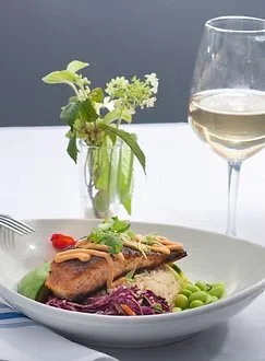 A plate of food with grilled meat, purple cabbage, green beans, and sauce, next to a glass of white wine on a white table.