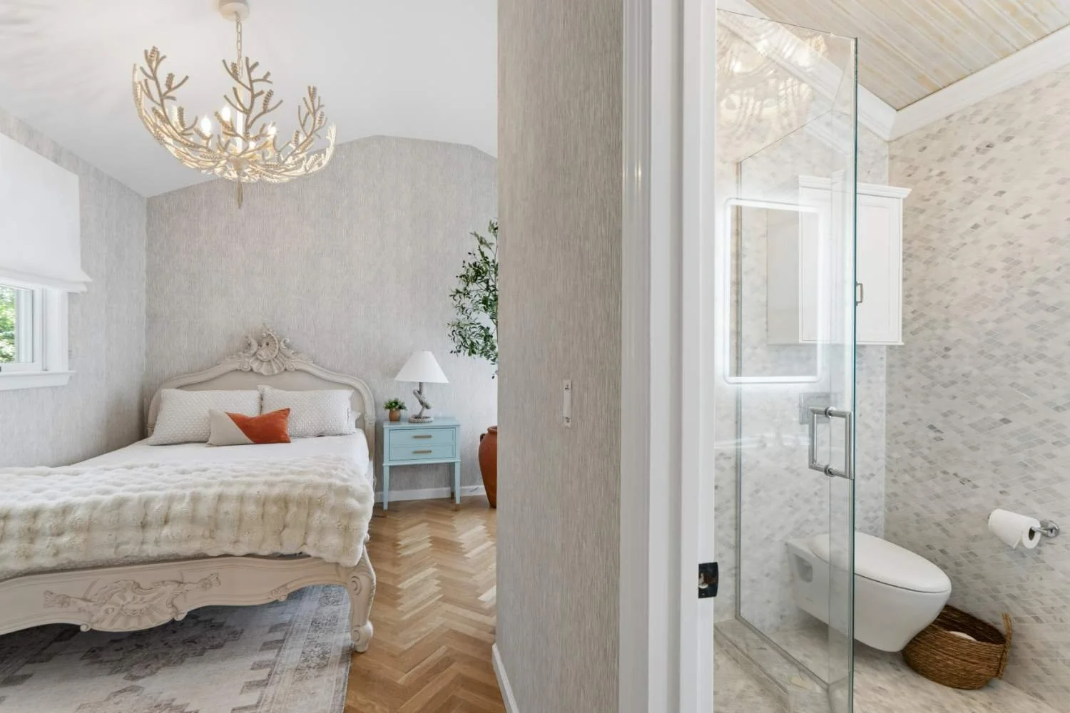A bedroom with a white ornate bed, a blue nightstand with a white lamp, a window with a shade, and neutral wall decor, seen through an open door from a bathroom with a glass shower, tiled walls, and a toilet.