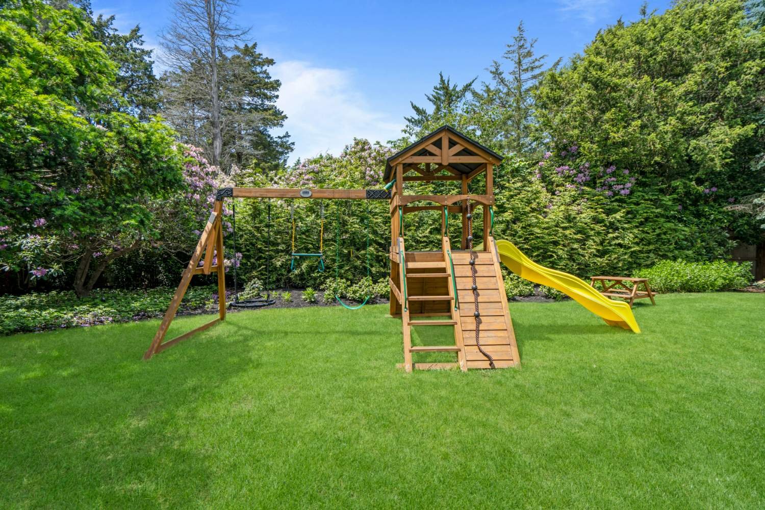 A backyard playground with a wooden swing set, slide, and climbing wall, surrounded by green grass and lush trees and shrubs.
