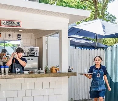 A young woman working at a small outdoor food stand with a white exterior, serving drinks. A young girl with a tray wears a blue apron and shirt. There is a large blue umbrella and a white fence in the background.