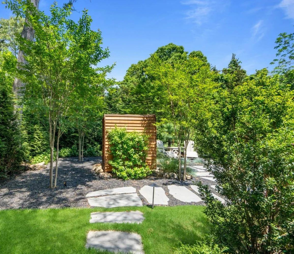A landscaped backyard with a stepping stone path, green grass, trees, shrubs, and outdoor furniture under a clear blue sky.