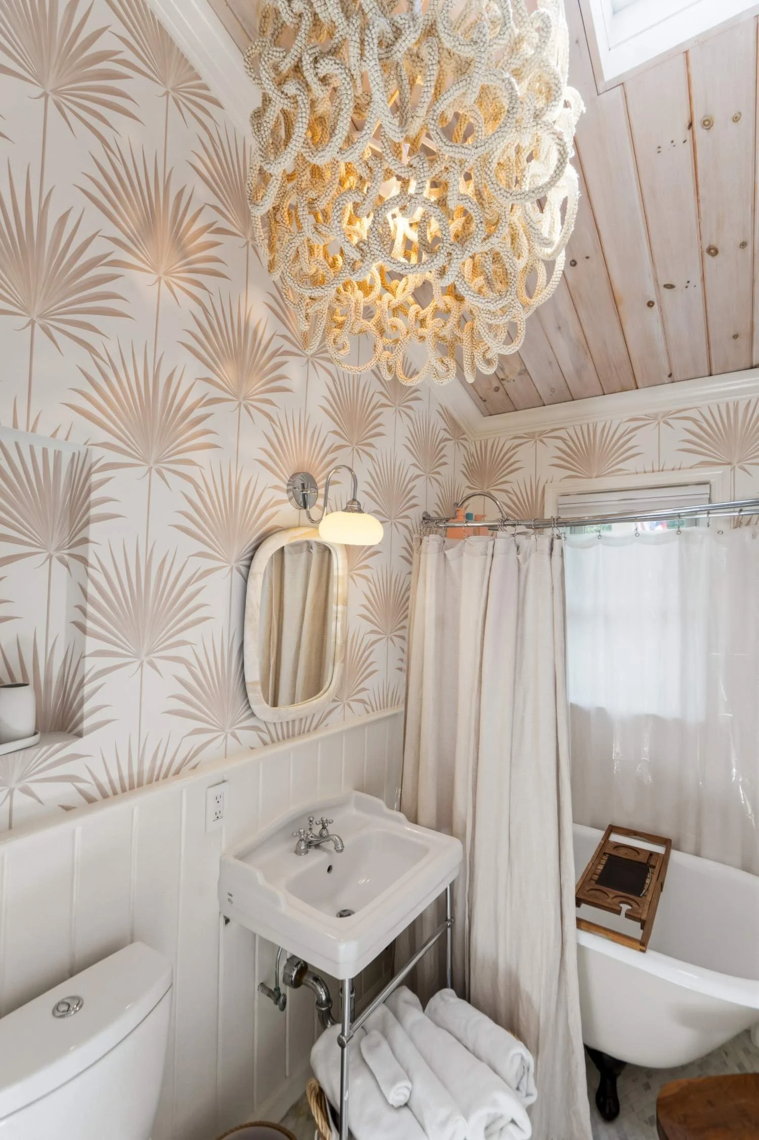 Bathroom with wallpaper that has a beige and white palm leaf pattern. Features include a white oval mirror, wall-mounted light fixture, small white sink, toilet, bathtub with a curtain, and a decorative wooden tray on a stool. A large chandelier made