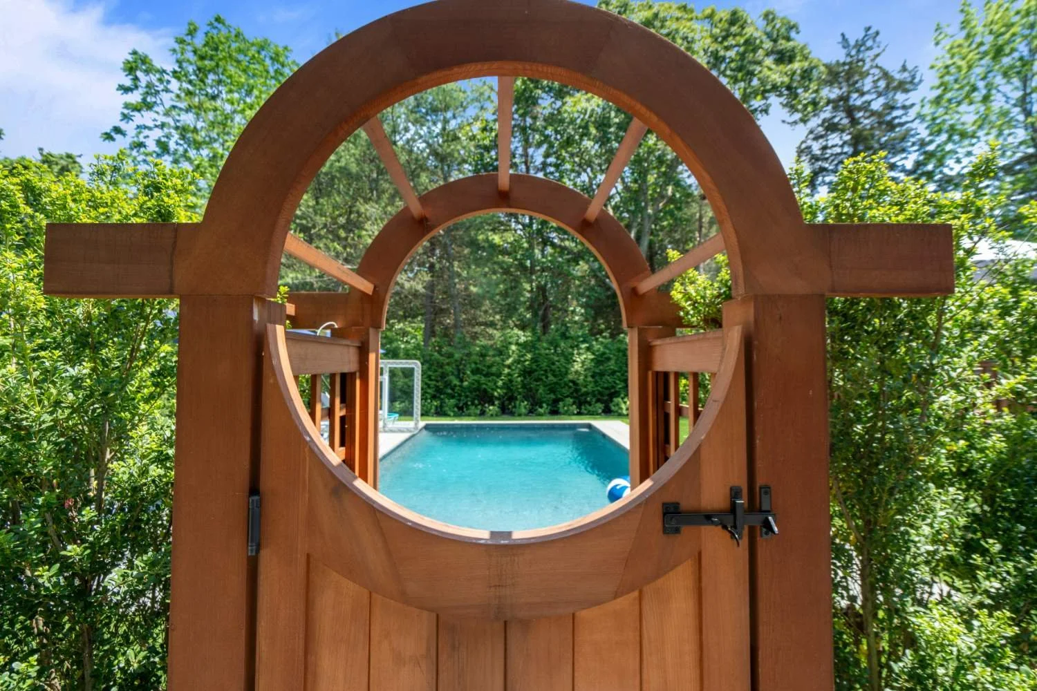 A wooden garden gate with a circular window, opening to a backyard with a swimming pool, surrounded by green trees and bushes.