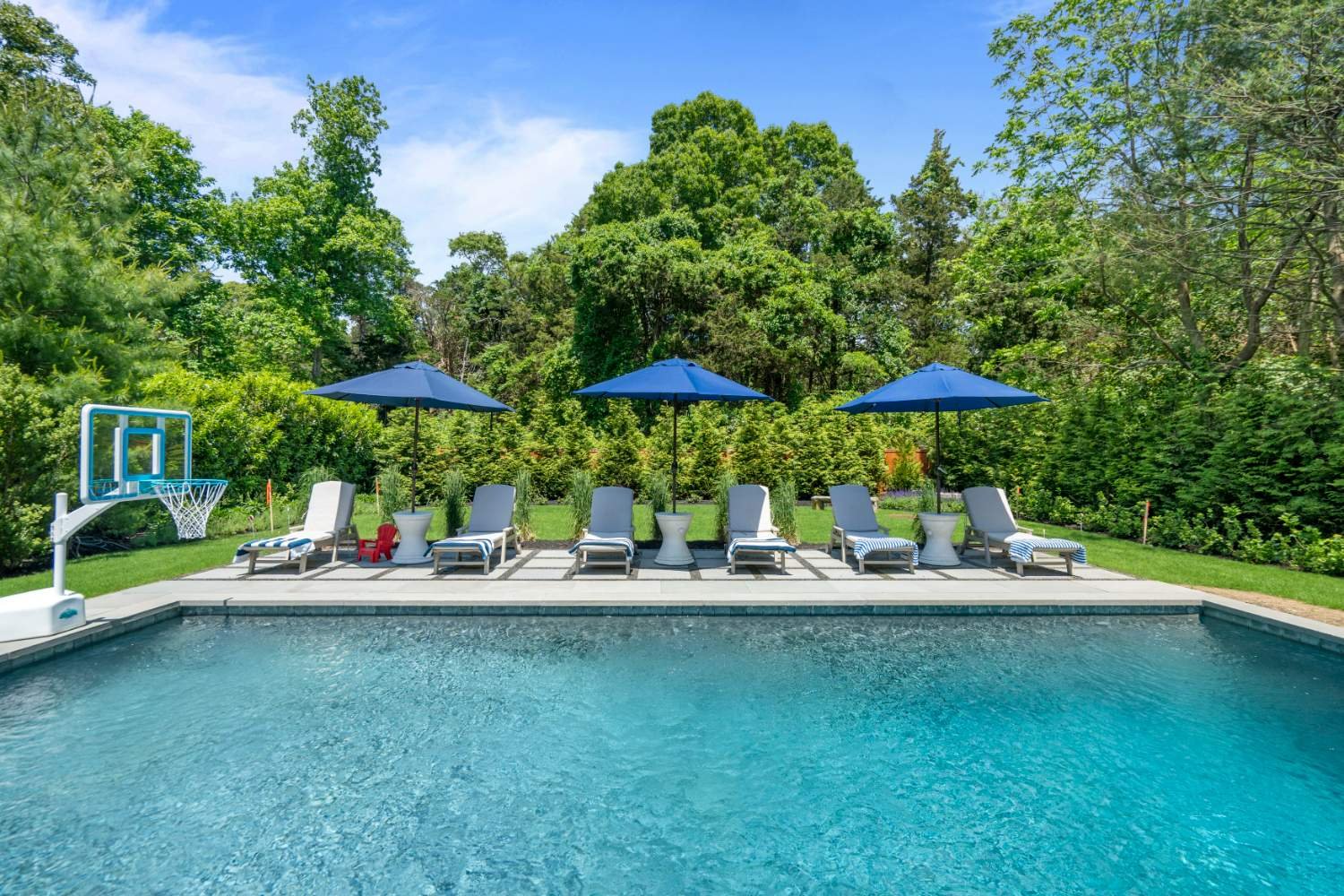 Swimming pool with blue water, surrounded by a tiled patio with six lounge chairs and three blue umbrellas. There are three white side tables between the chairs. In the background, dense green trees and bushes enclose the area, with a clear blue sky 