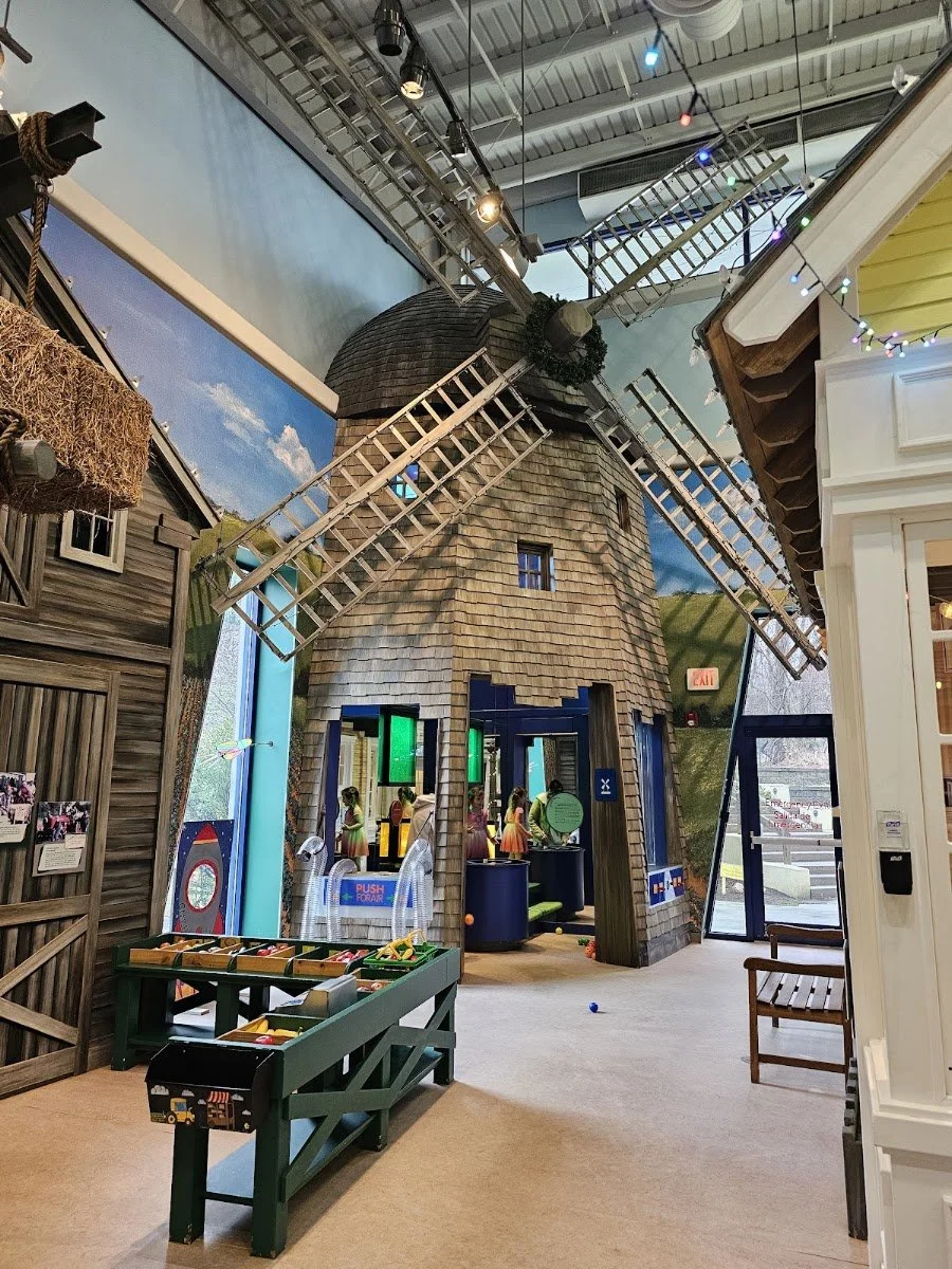Indoor play area with a large, model windmill with sails, a raised wooden house with hay bales, arcade games, and children playing.