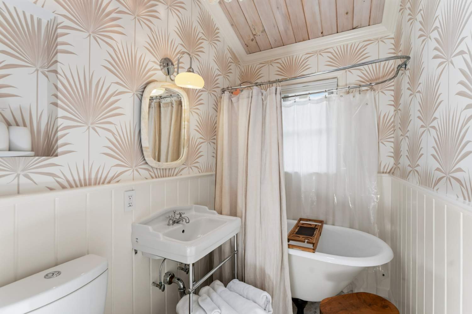 A small bathroom with tropical leaf-patterned wallpaper, a white toilet, a small porcelain sink, an oval mirror, a wall-mounted light fixture, a shower with a curtain, a clawfoot bathtub with a wooden tray, and a small wooden stool.