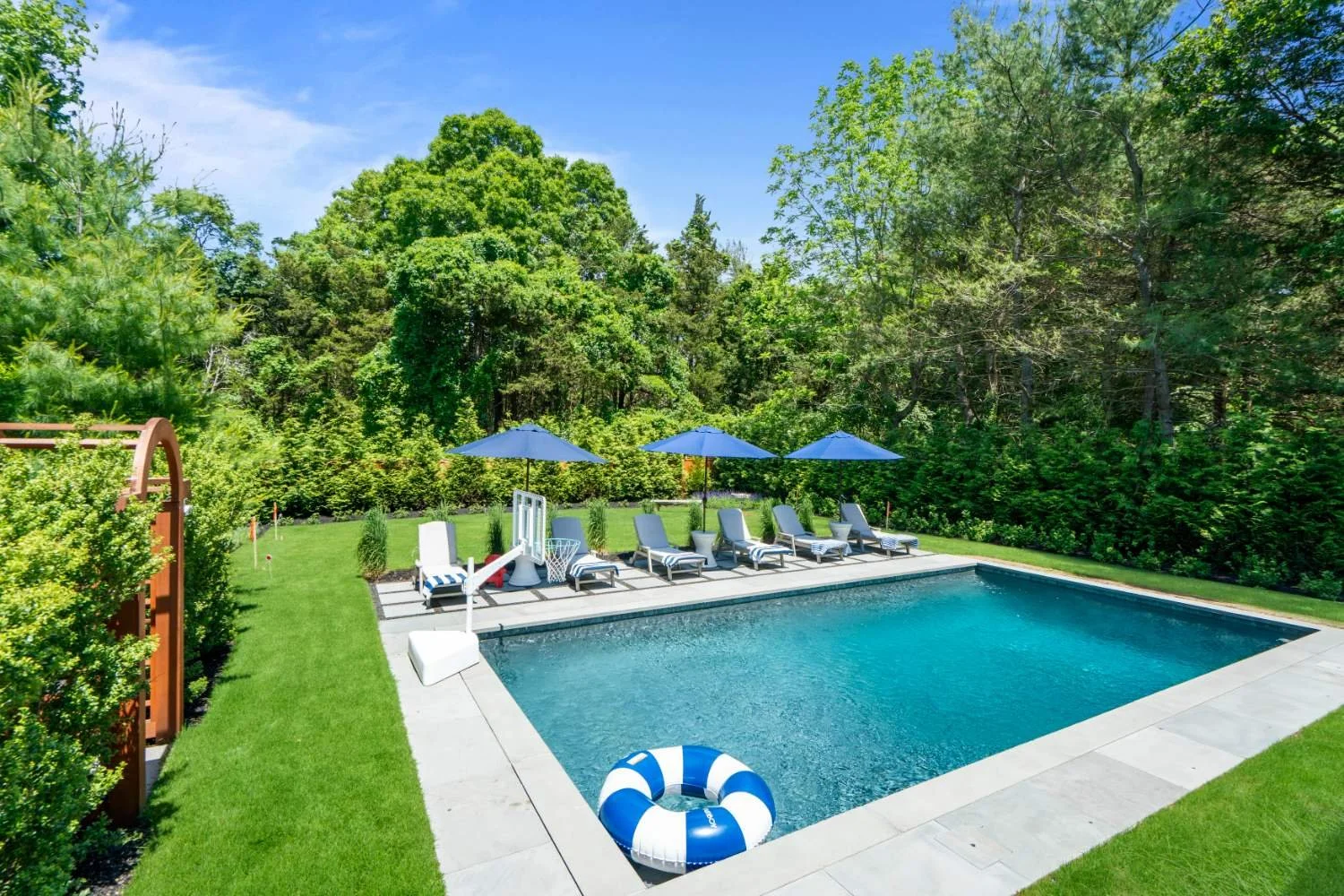 Residential backyard with a rectangular swimming pool, surrounded by green grass, lush trees, and bushes. There are five lounge chairs with blue umbrellas, a basketball hoop, and a life ring float in the pool. A wooden arbor is visible on the left si