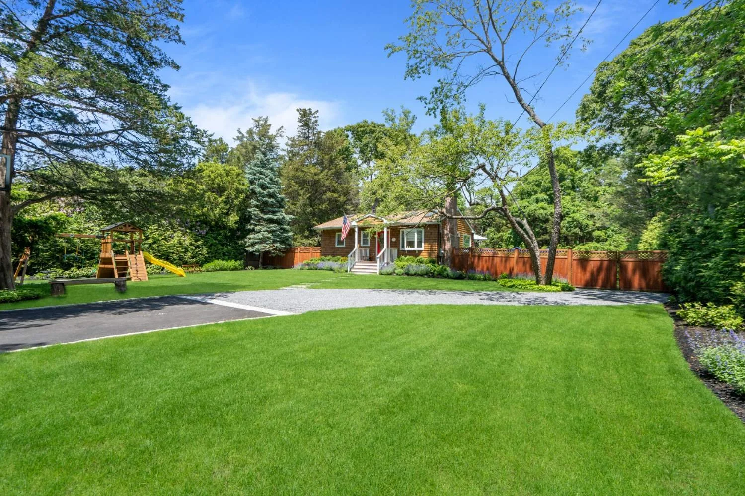 A well-maintained backyard with green grass, a children's playset with a slide, a paved driveway, and a house with wooden siding surrounded by trees and shrubs, under a clear blue sky.
