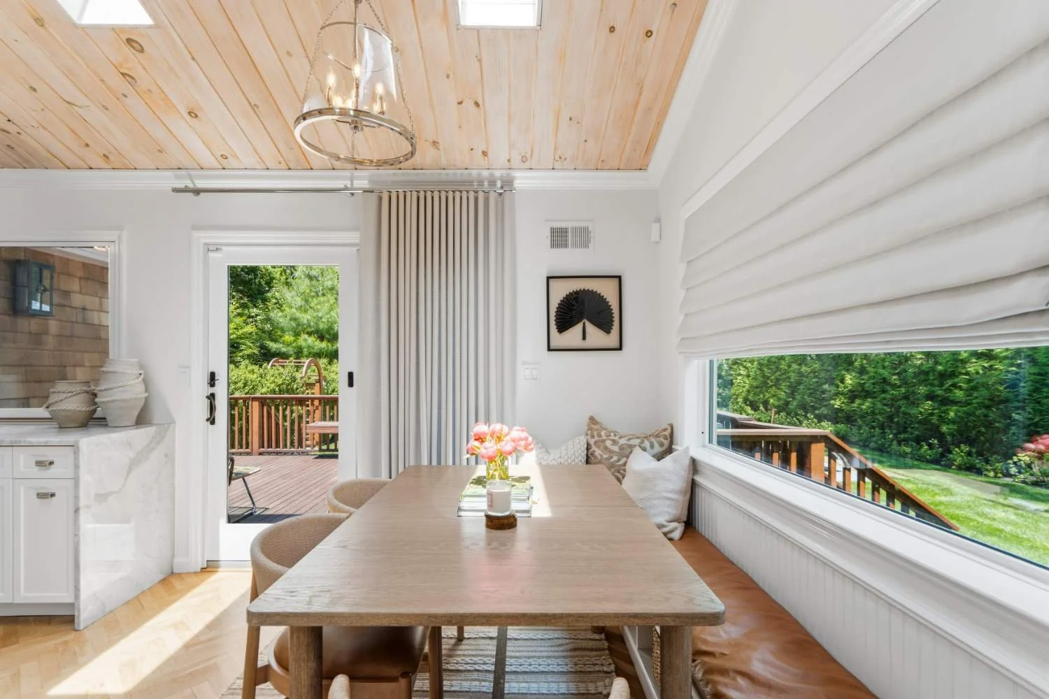 A dining area with a wooden table, beige chairs, and a window seat with pillows. Outside are a deck and green trees. The ceiling is wood, and the room has white walls and curtains.