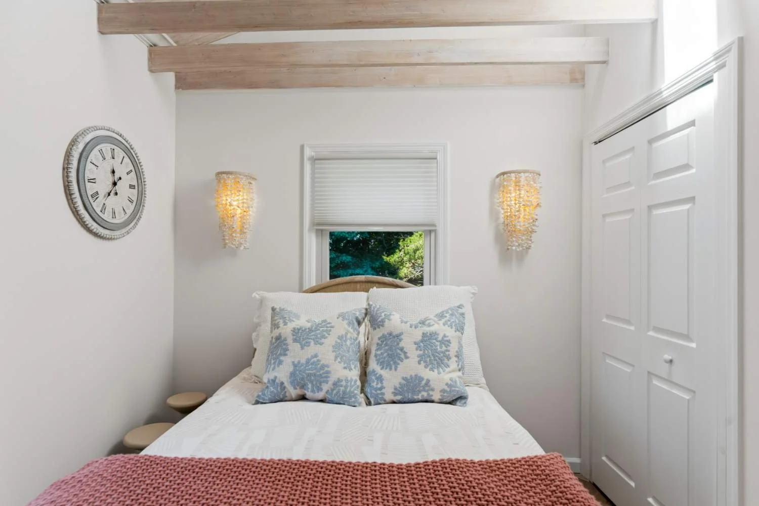 A cozy bedroom with white walls and a wooden ceiling. The bed has white bedding, two decorative pillows with blue leaf patterns, and a pink knitted blanket. There are two wall-mounted lamps with shell-like shades, a window behind the bed showing gree