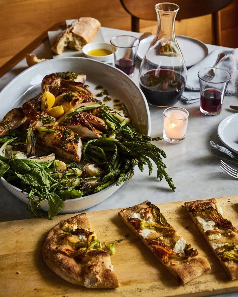 A table set with a roasted chicken, grilled vegetables, pizza slices, a carafe of red wine, glasses of wine, bread, and a lit candle.