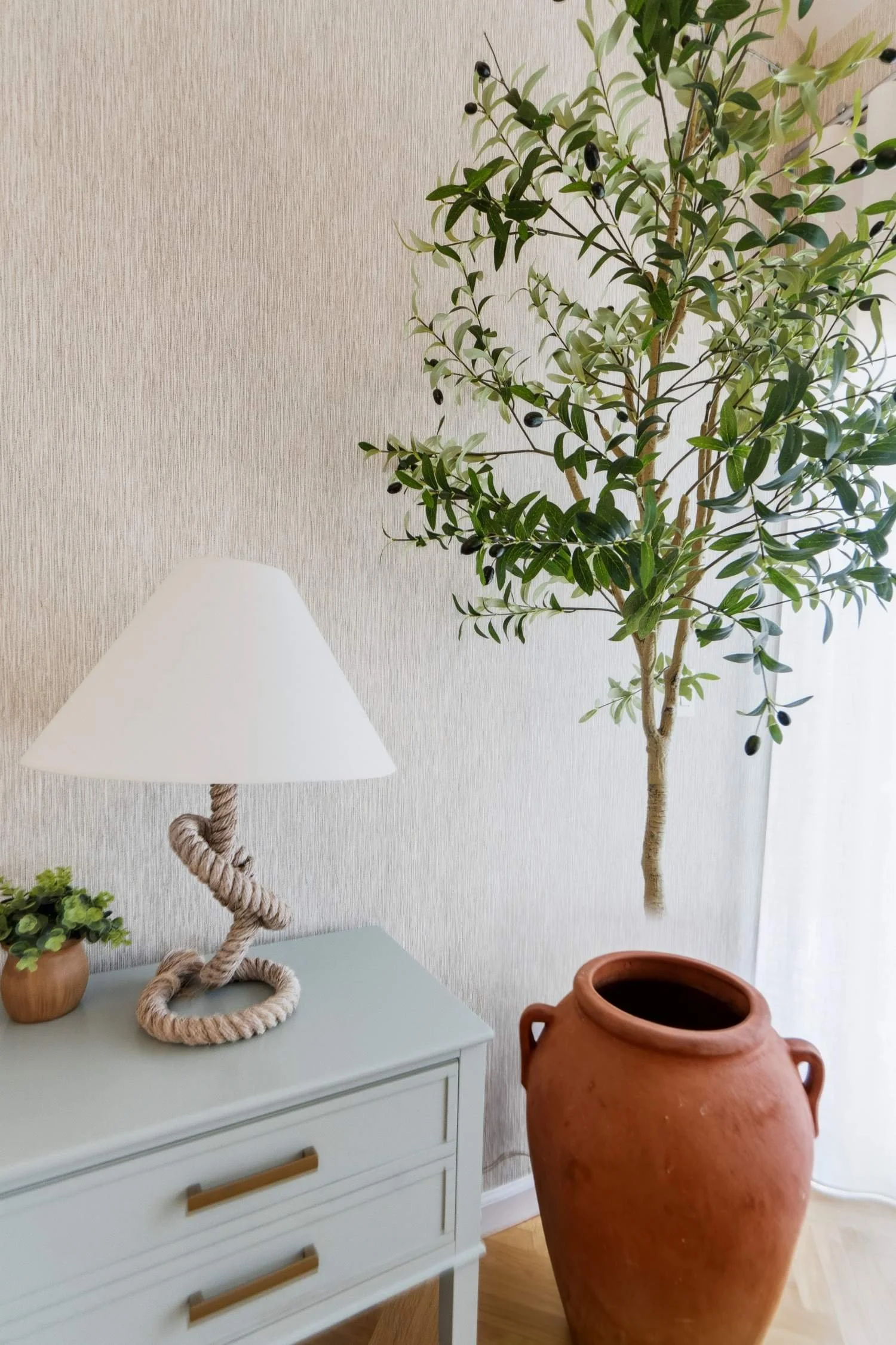 A living room corner with a white dresser, a table lamp with a rope base, a small potted plant, a large terracotta vase, and a tall indoor tree with green leaves and black berries, against a beige textured wall.