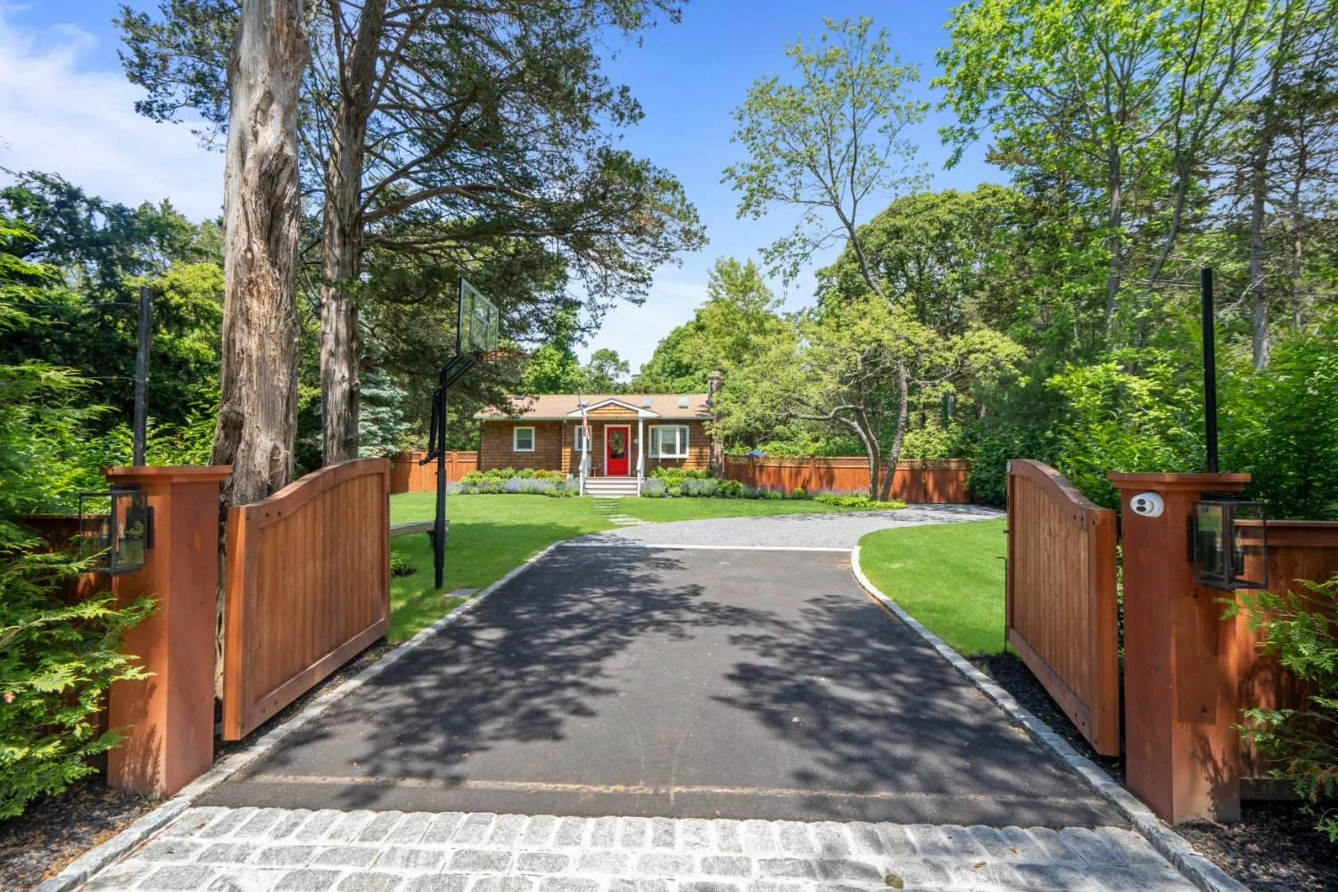 A view of a house with a red front door, surrounded by green trees and a well-maintained garden, seen from an open wooden gate, with a driveway leading up to the house on a sunny day.