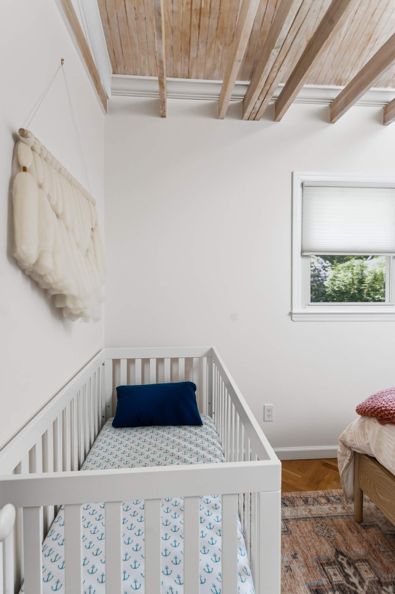 A baby crib with a navy blue pillow and a mattress covered in white sheets with blue anchors pattern. The room has a white wall, a window with a closed blind, a wall hanging made of soft, cream-colored fabric, wooden beams on the ceiling, and a patte