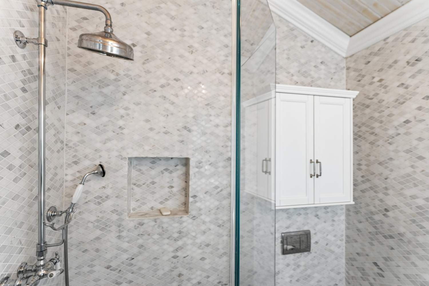 A bathroom with a tiled shower area featuring a chrome showerhead, a built-in niche with soap, a handheld shower wand, and a white cabinet above a hidden toilet tank.