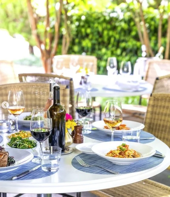 Outdoor dining table set with plates of pasta, salad, glasses of wine, and wine bottles, surrounded by wicker chairs with greenery in the background.