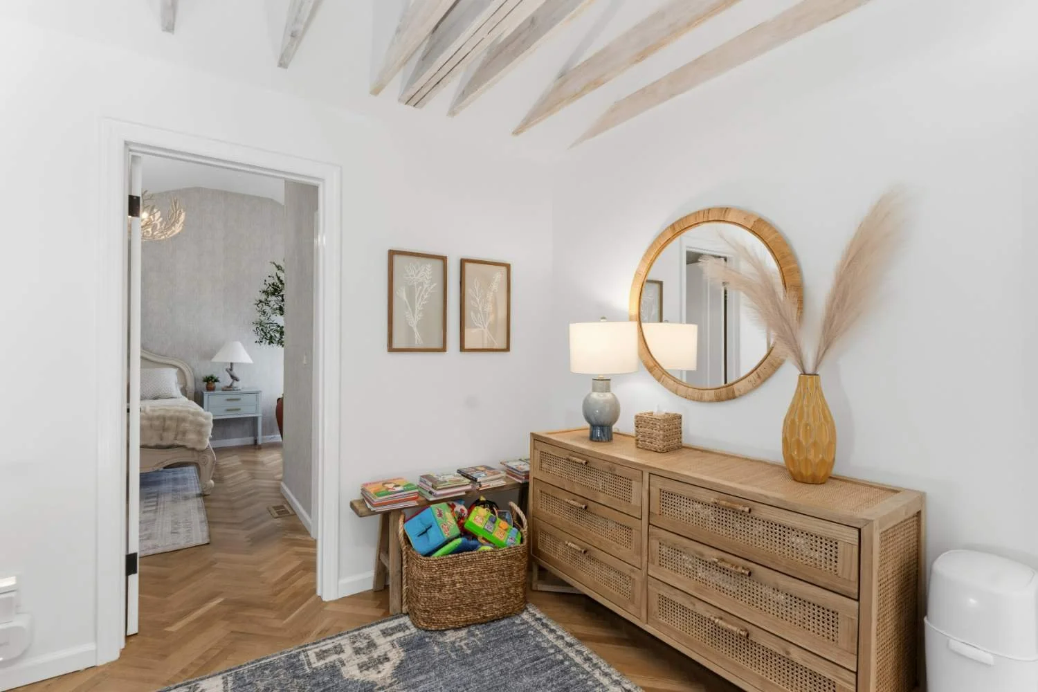 Living room corner with wooden dresser, round mirror, table lamp, basket of children's toys, framed botanical art, and beige decorative vase with pampas grass, hardwood floor, and part of an adjacent bedroom visible through doorway.