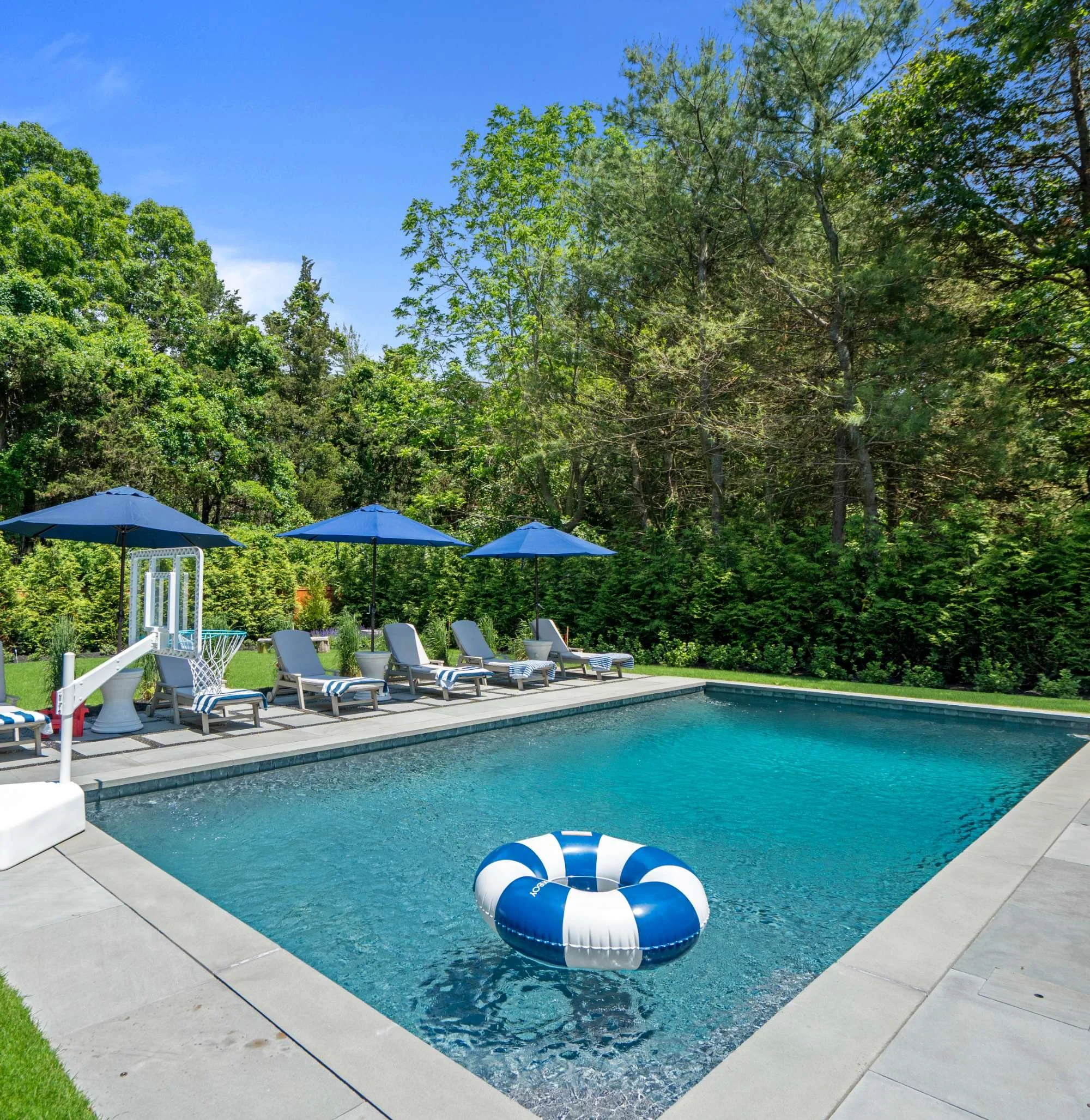An outdoor swimming pool with yellow lounge chairs and blue umbrellas in a backyard surrounded by green trees and bushes, with a blue sky overhead.