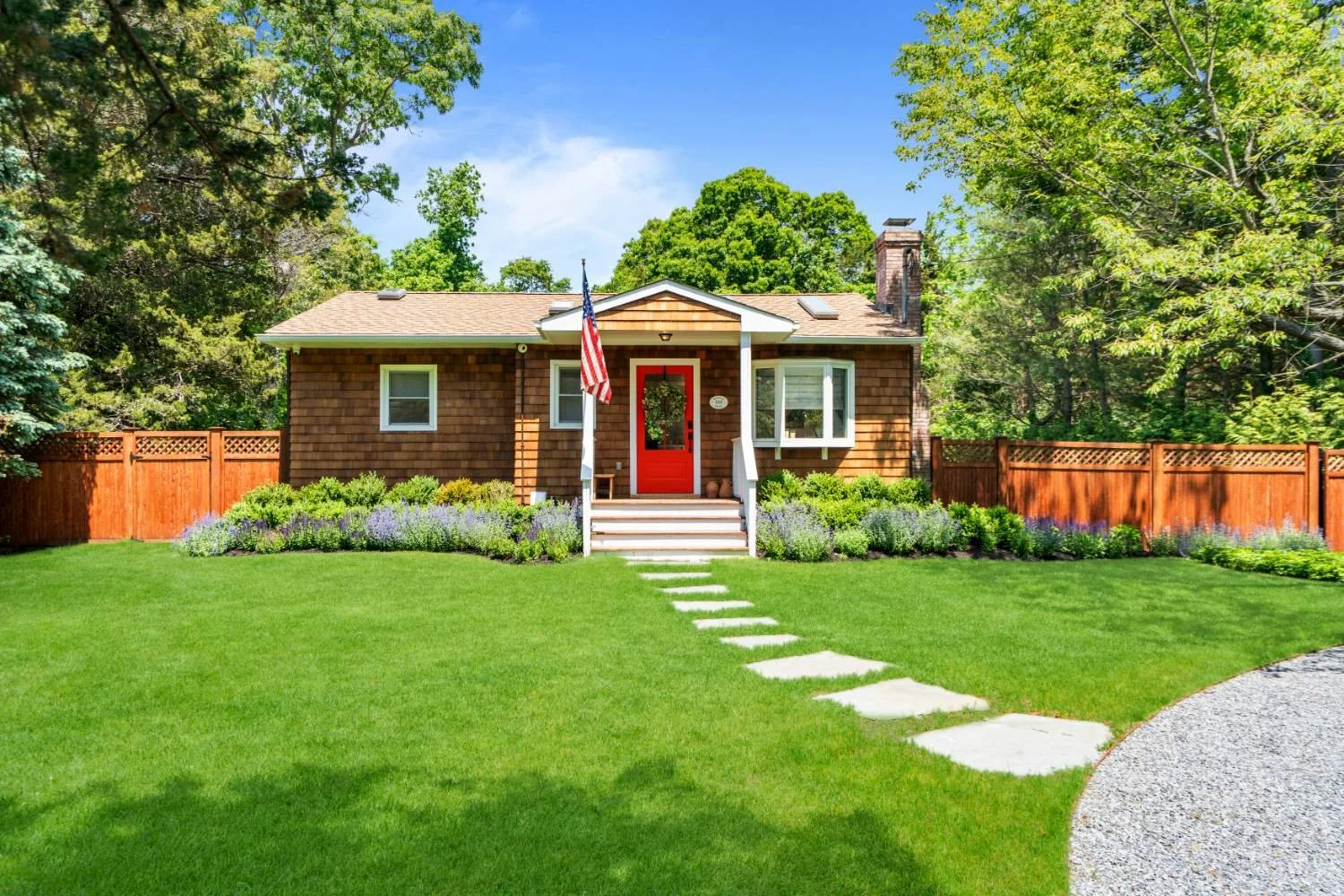Front view of a house with a red door, surrounded by a green lawn, garden, and trees, and enclosed by a wooden fence.